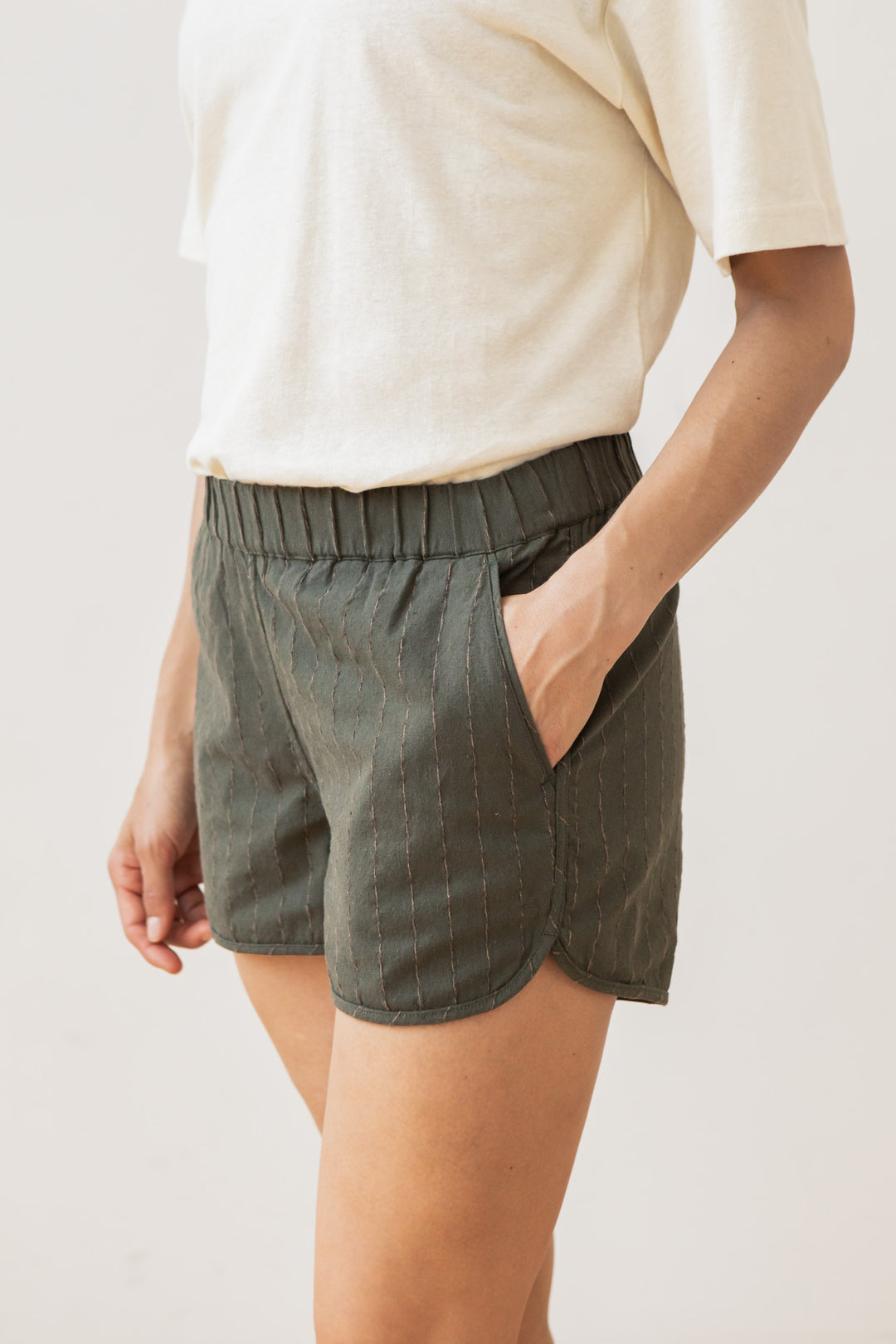 A person wears Bridge & Burn's Luca Track Short in Olive Stripe, styled with side pockets and subtle vertical stitching, paired with a short-sleeved off-white shirt, standing against a plain background.
