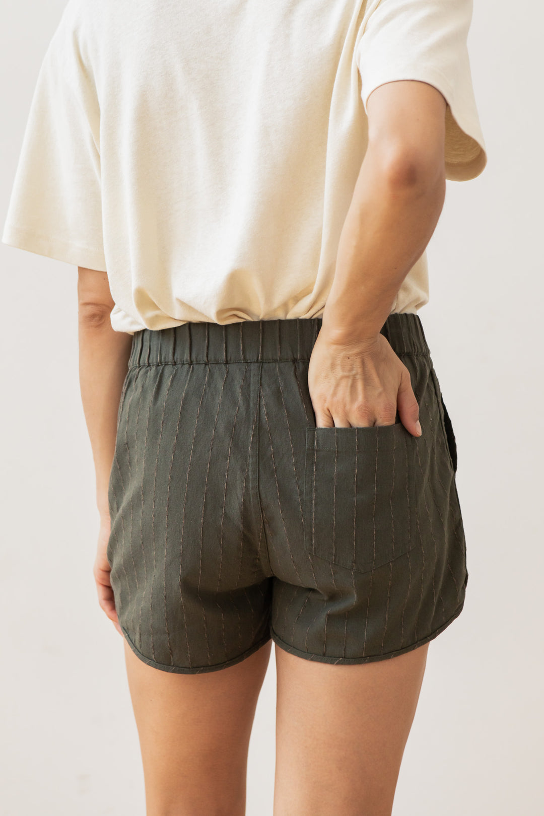 A person is shown from the back wearing Bridge & Burn’s Luca Track Short in Olive Stripe with a loose white t-shirt, one hand placed in the back pocket of the shorts.