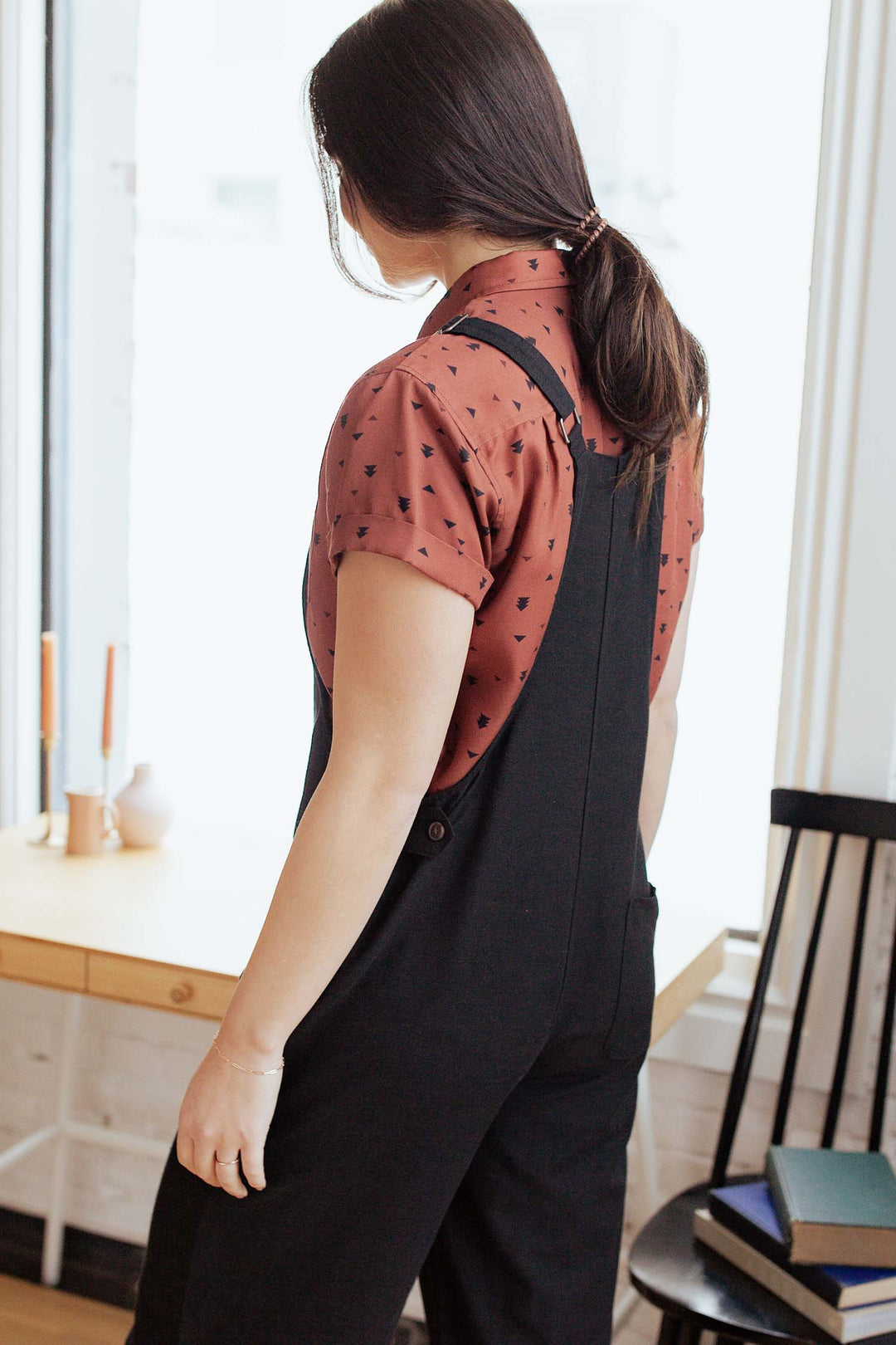 A person with long dark hair in a ponytail stands indoors, wearing the Bridge & Burn Edie Overall in Black over a rust-patterned shirt. Nearby are a wooden chair and table with candles.