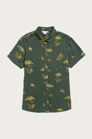 The Bridge & Burn Lana Shirt in Woodland Bloom features a relaxed fit, short sleeves, button-up front, and a dark green base with yellow and white florals. Made from an eco-friendly cotton alternative with a chest pocket.