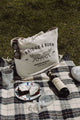 A picnic on grass with a plaid blanket, sunglasses, two crystal cups, a wine bottle, a plate with two brownies, part of a sneaker, and the Bridge & Burn Fox Tote in Natural.