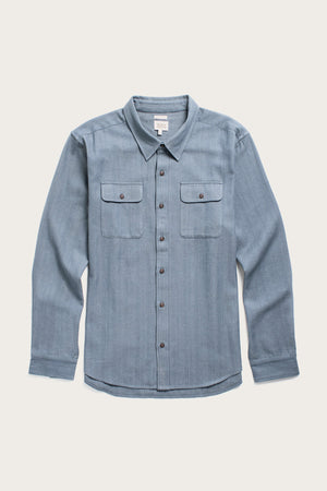 The Bridge & Burn Bedford Shirt in denim herringbone is a light blue, long-sleeve organic cotton shirt with a collar and two button-flap chest pockets, shown laid flat on a plain white background.