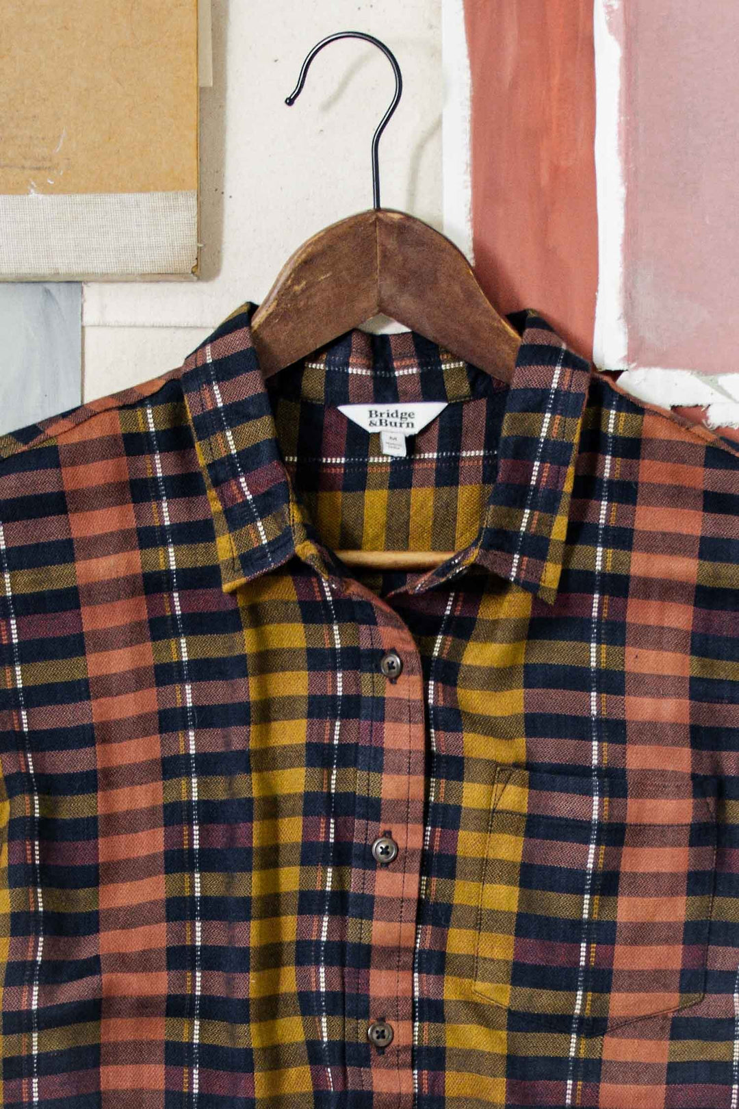 The Bridge & Burn Bea Shirt in Alpenglow Stripe, an eco-friendly short-sleeve button-down, features mustard yellow, rust, and black stripes and hangs on a wooden hanger against neutral and pinkish panels.