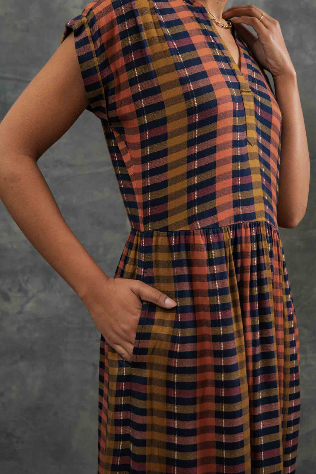 A person models the Bridge & Burn Ira Dress in Alpenglow Stripe—an eco-friendly, short-sleeved plaid pullover with orange, navy, and yellow stripes. One hand is in a pocket; the other touches their neck. Face not visible; background is gray.