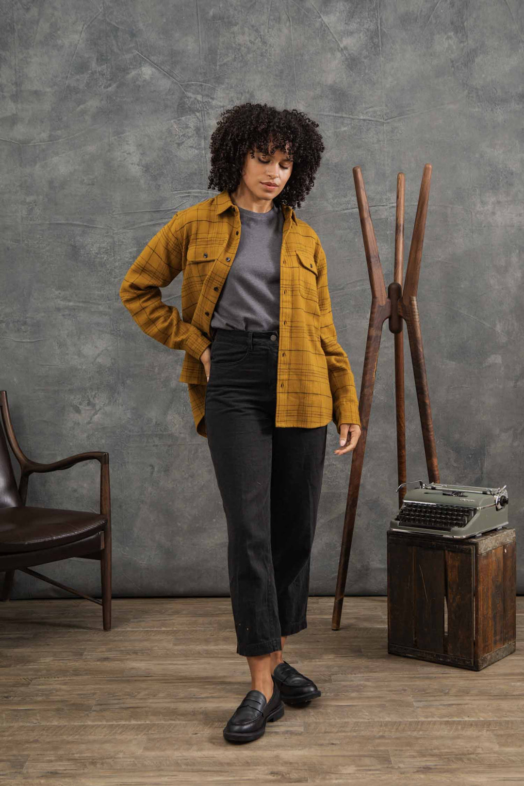 A woman with curly hair wears the Bridge & Burn Lenon Shirt in Golden Horizon Plaid over a gray t-shirt, black pants, and black loafers. She stands indoors near a wooden chair, vintage typewriter, and coat rack.
