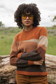 A person with curly hair and large yellow sunglasses stands outdoors, arms crossed, wearing the Bridge & Burn Petra Sweater in Sandstone, featuring earthy stripes, in front of a fallen log and grassy field.