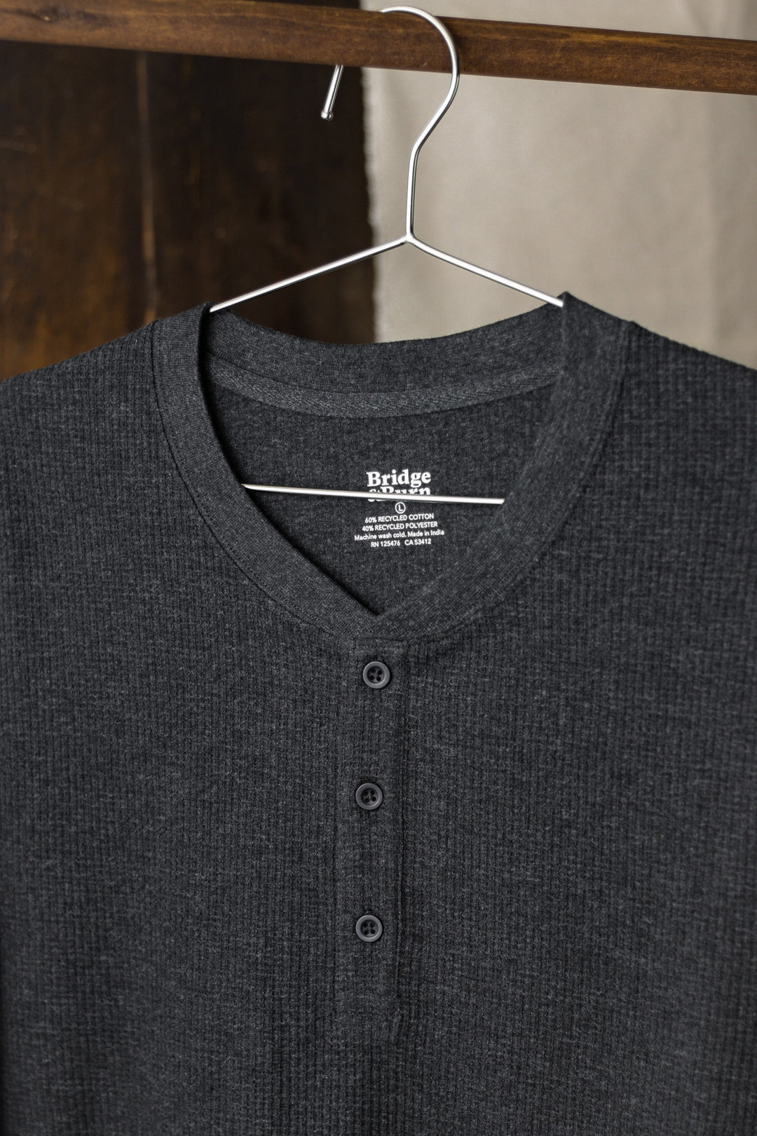The Bridge & Burn Women's Recycled Waffle Henley in Charcoal Melange features recycled fibers, a ribbed long-sleeve waffle-knit design, and three front buttons, shown hanging on a thin metal hanger against a wooden and beige background.
