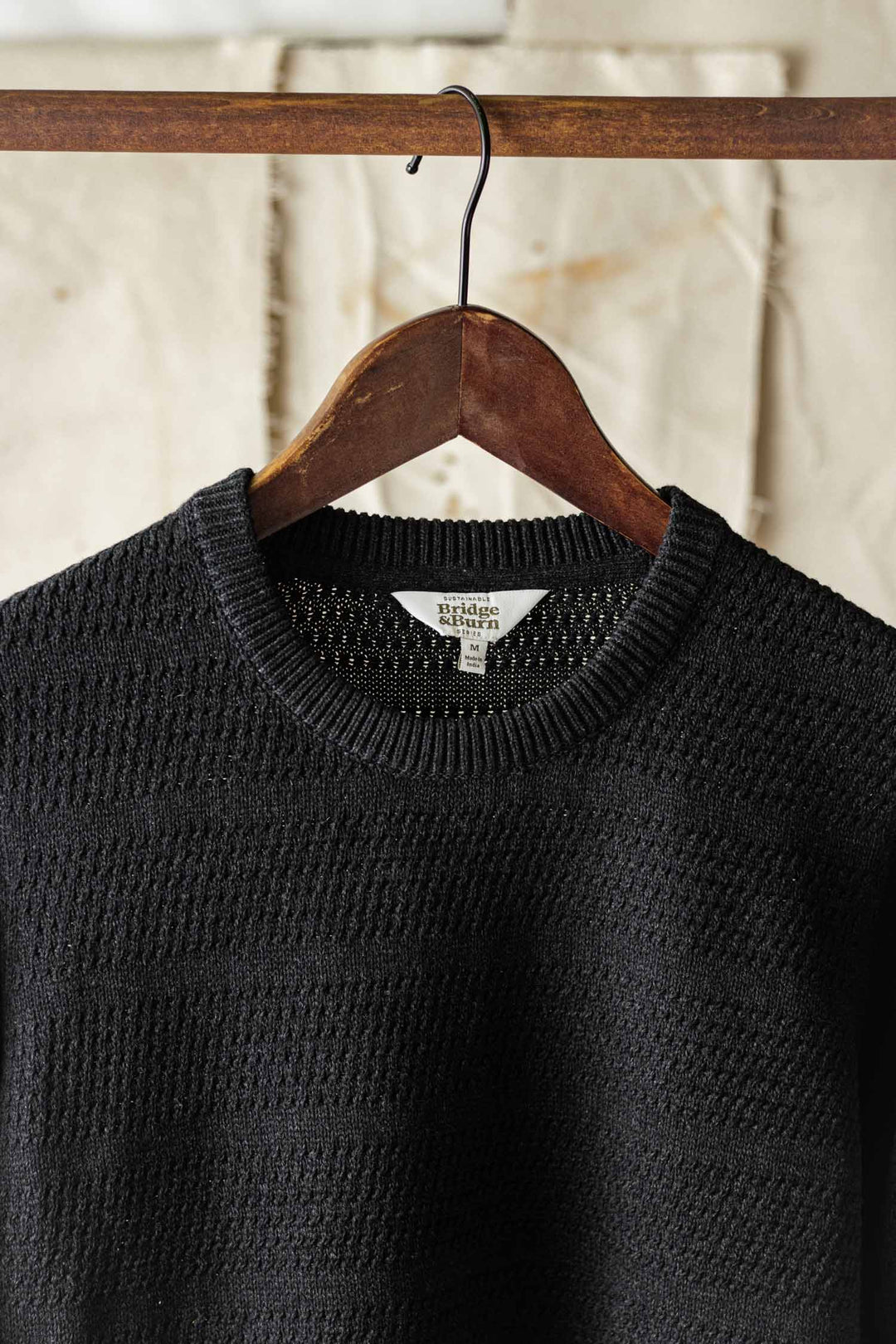 The Bridge & Burn Calder Sweater in Midnight, featuring a textured knit design, hangs on a wooden hanger from a rod against a neutral fabric backdrop.
