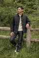 A man stands outdoors by a wooden fence, wearing the Bridge & Burn Holman Quilted Jacket in Black Waxed Canvas, paired with a white sweater, dark jeans, and black sneakers. Green grass and trees create a relaxed natural setting.