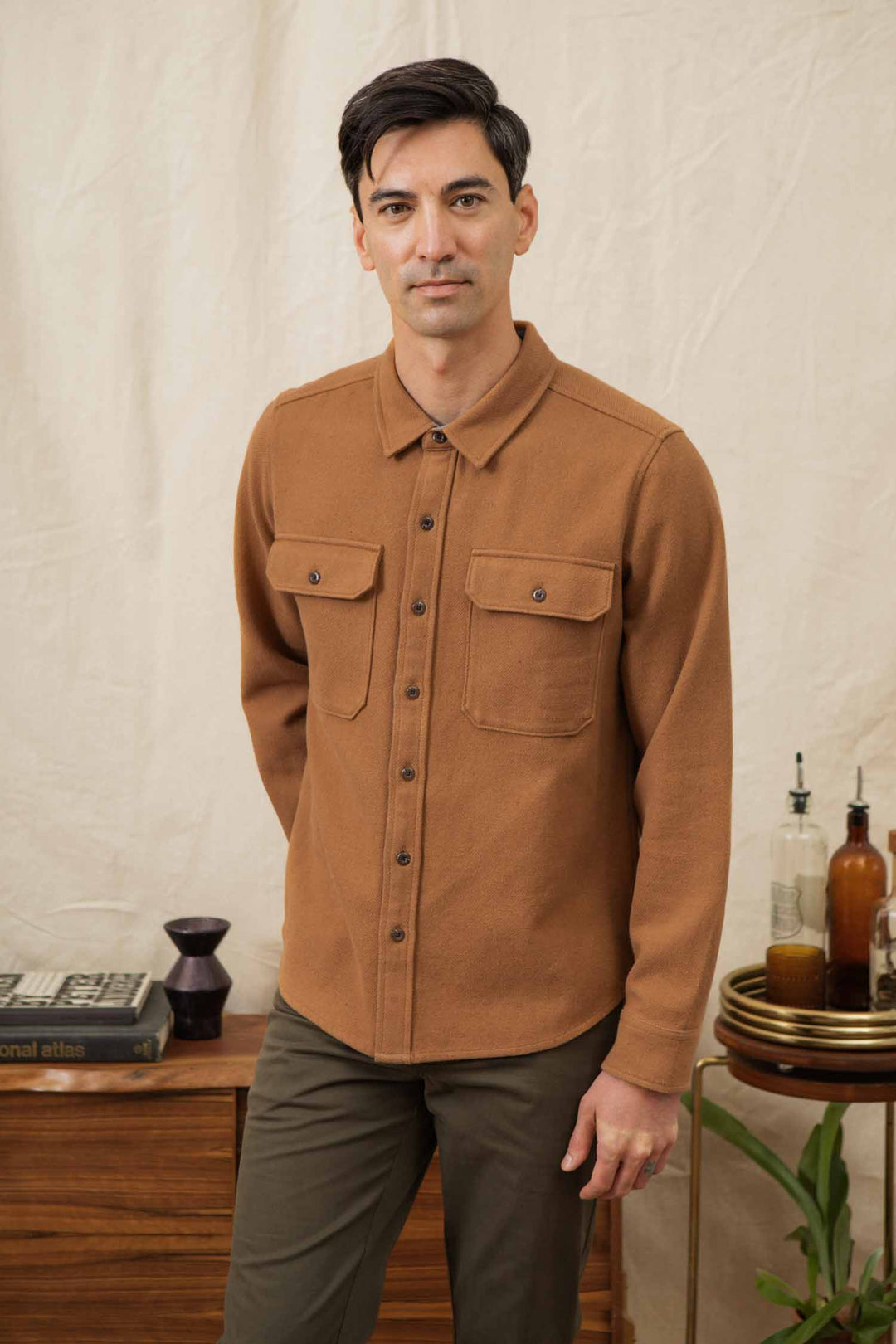 A man with short dark hair wears the Bridge & Burn Pointer Overshirt in Camel with chest pockets and olive pants, standing indoors near a wooden cabinet with books, vases, and bottles on it against a beige backdrop.