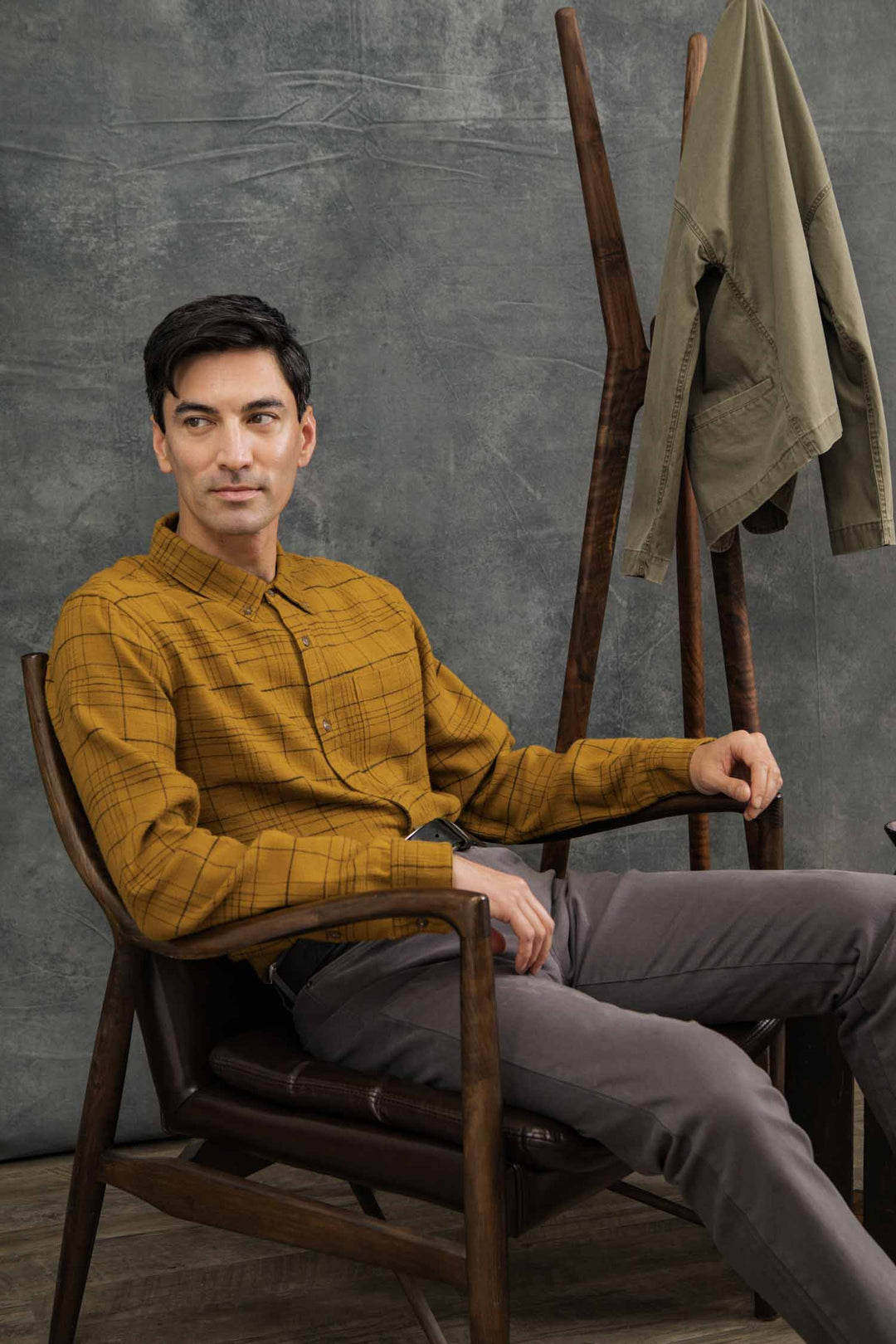 A man with short dark hair wears the Bridge & Burn Sutton Slim Shirt in Golden Horizon Plaid and gray pants, sitting in a wooden chair. Behind him, an olive green jacket hangs on a coat rack against a gray textured wall.