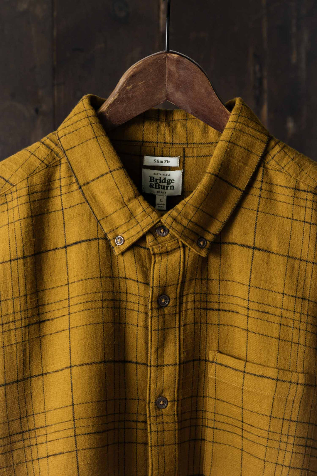 A Sutton Slim Shirt / Golden Horizon Plaid by Bridge & Burn hangs on a wooden hanger against a dark wood background, featuring a mustard yellow slim fit flannel with a minimalist black windowpane plaid.