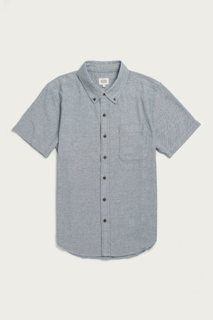 The Bridge & Burn Bryce Shirt in Denim Twill is a light blue, short sleeve button-down with a collar and chest pocket, made from recycled cotton for sustainability, shown on a white background.