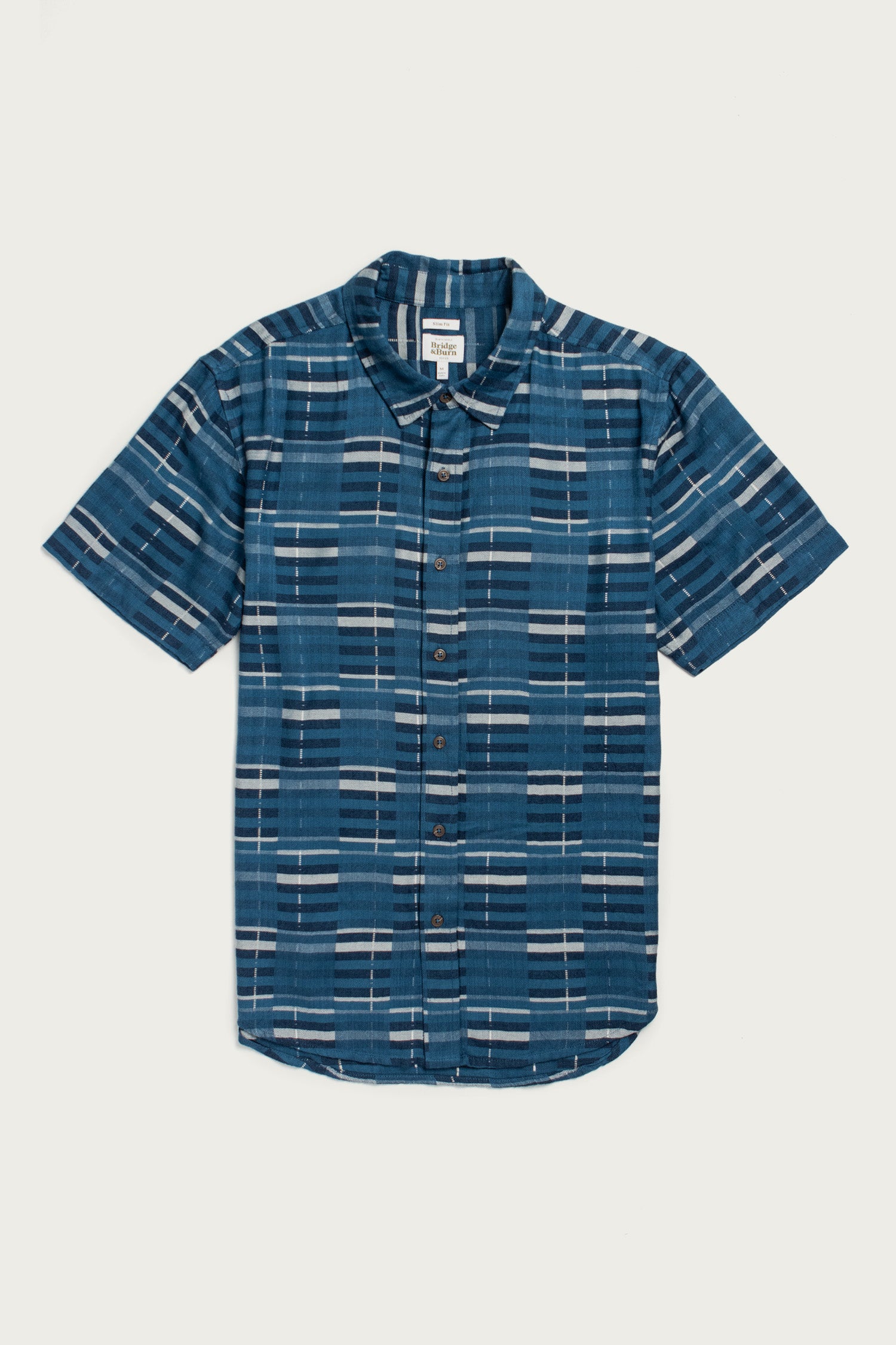 The Emerson Slim Shirt by Bridge & Burn features short sleeves, button-up front, and a blue base with white and dark blue geometric stripes, shown laid flat on a white background.