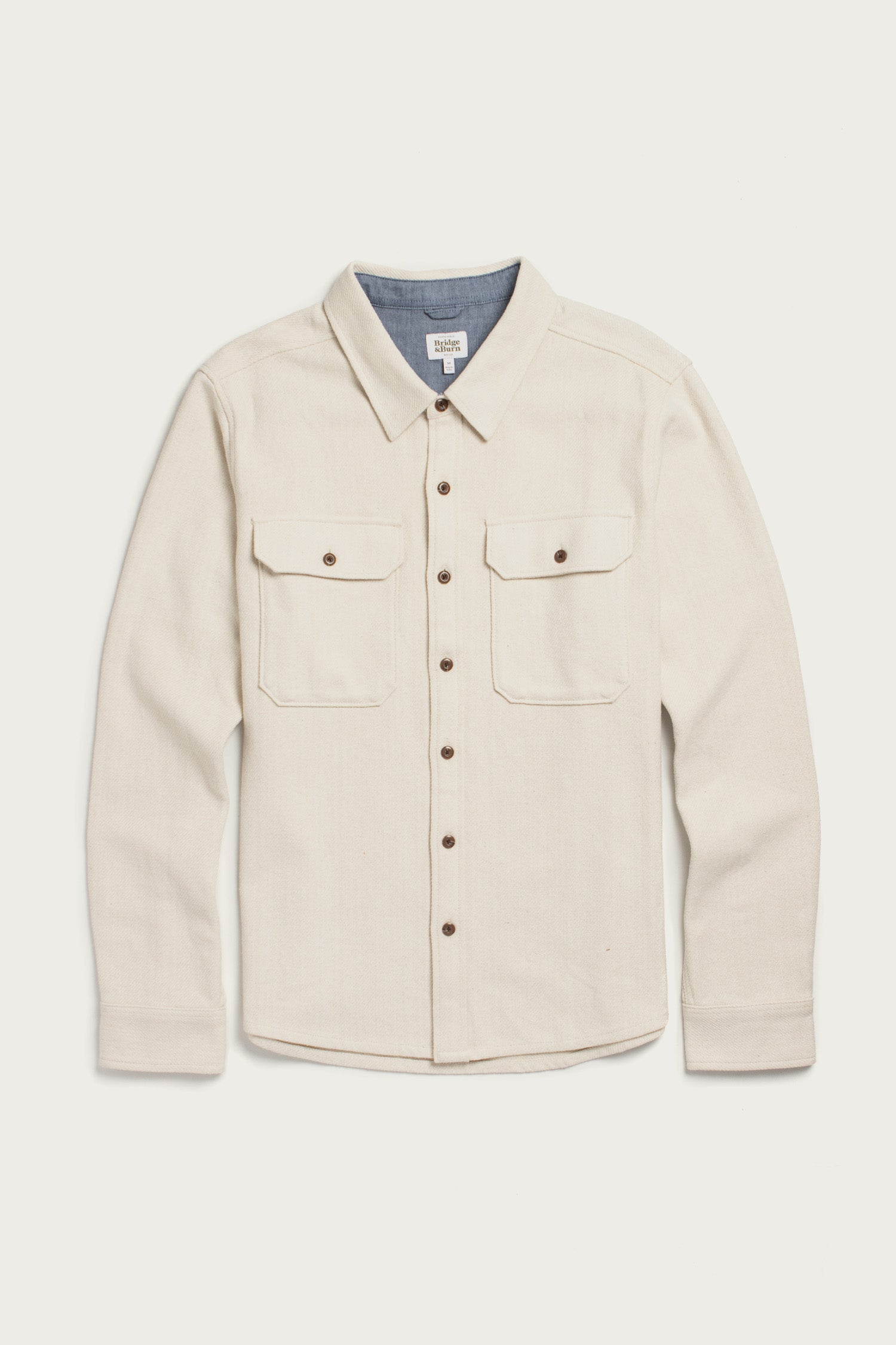 The Bridge & Burn Pointer Overshirt in Ivory Twill is a long-sleeve overshirt with two chest pockets, brown buttons, and a pointed collar, made from recycled fibers and shown laid flat on a white background.