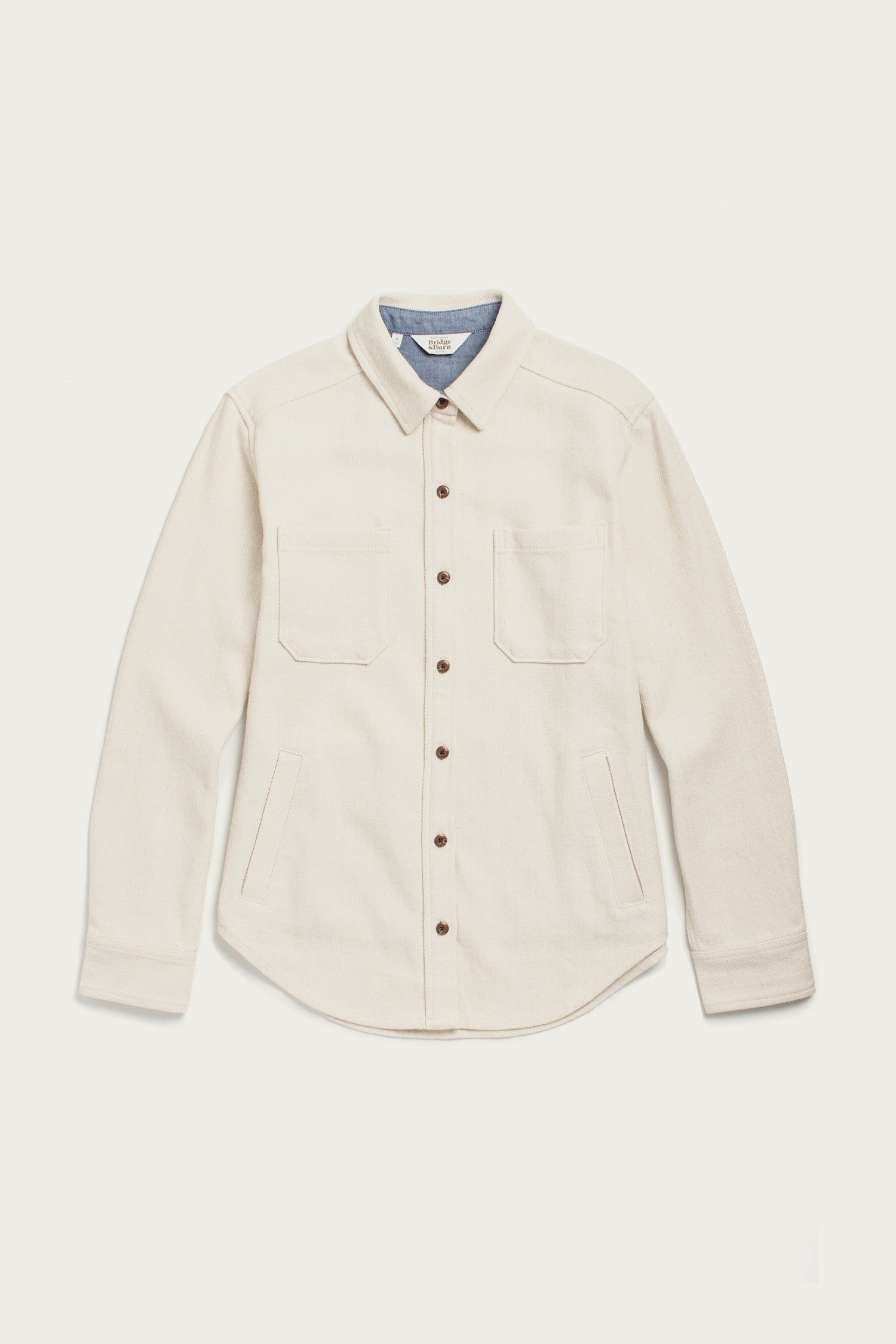 The Bridge & Burn Dawson Overshirt in Ivory Twill is a cream-colored, recycled cotton button-up with brown buttons, a light blue inner collar lining, two chest pockets, side pockets, and long sleeves. Laid flat on a white background.