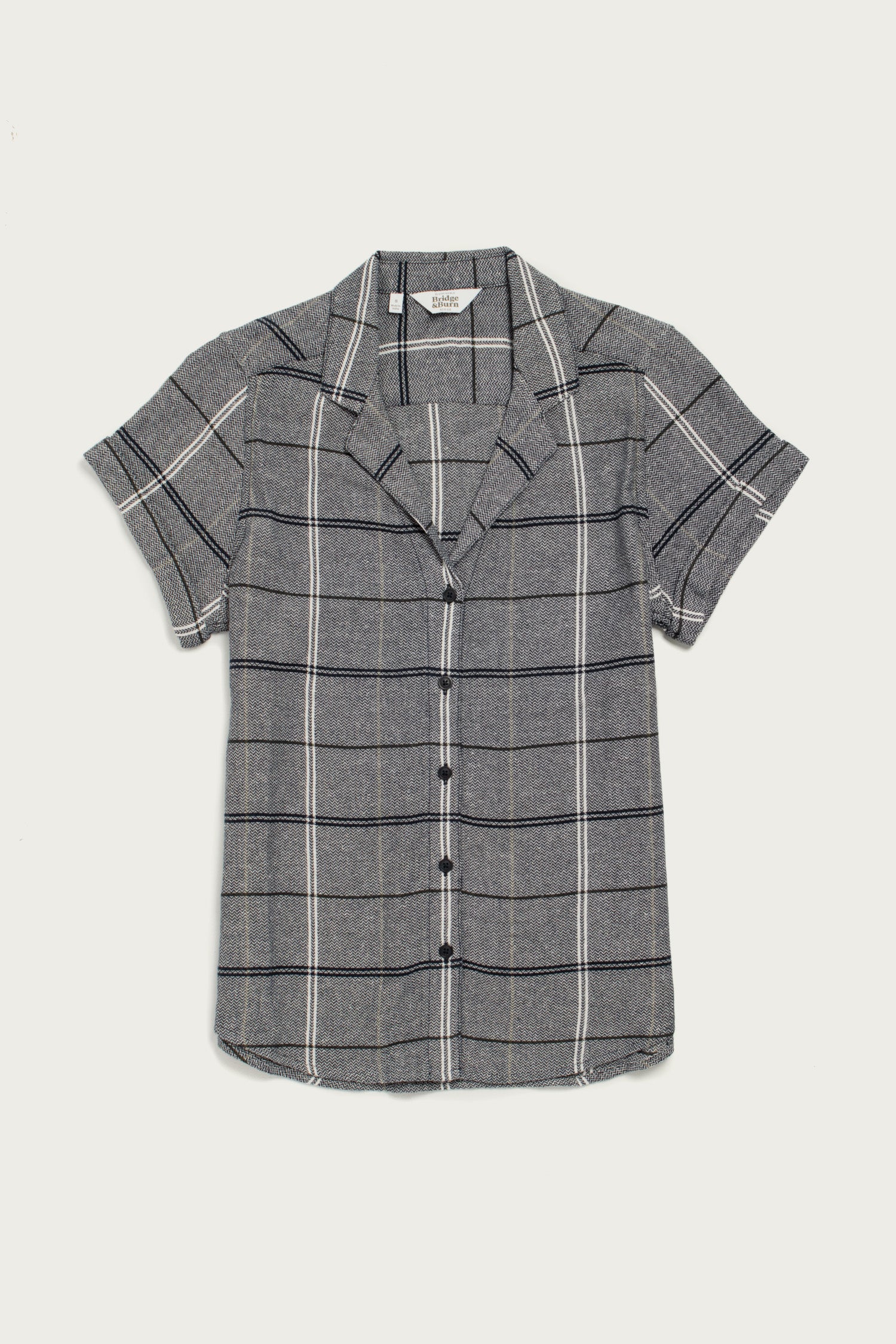 The Bridge & Burn Innes Shirt in Overcast Grid is a relaxed fit, short-sleeve button-up made from recycled cotton with a gray base and white and black plaid, shown on a plain white background.