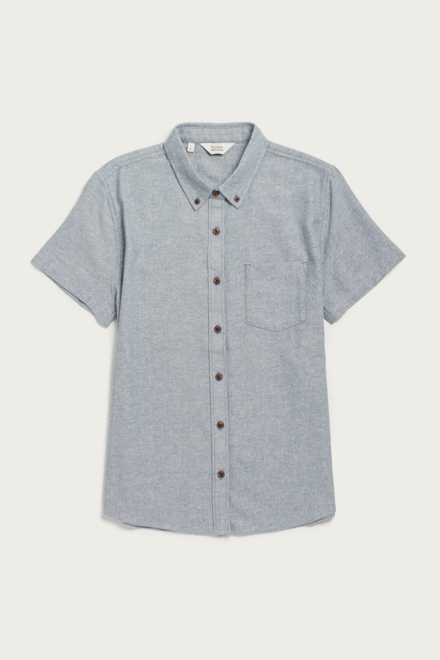 The Bridge & Burn Juniper Shirt in denim twill features short sleeves, a button-down collar, chest pocket, relaxed fit, and is crafted from recycled cotton. Shown here laid flat on a white background.