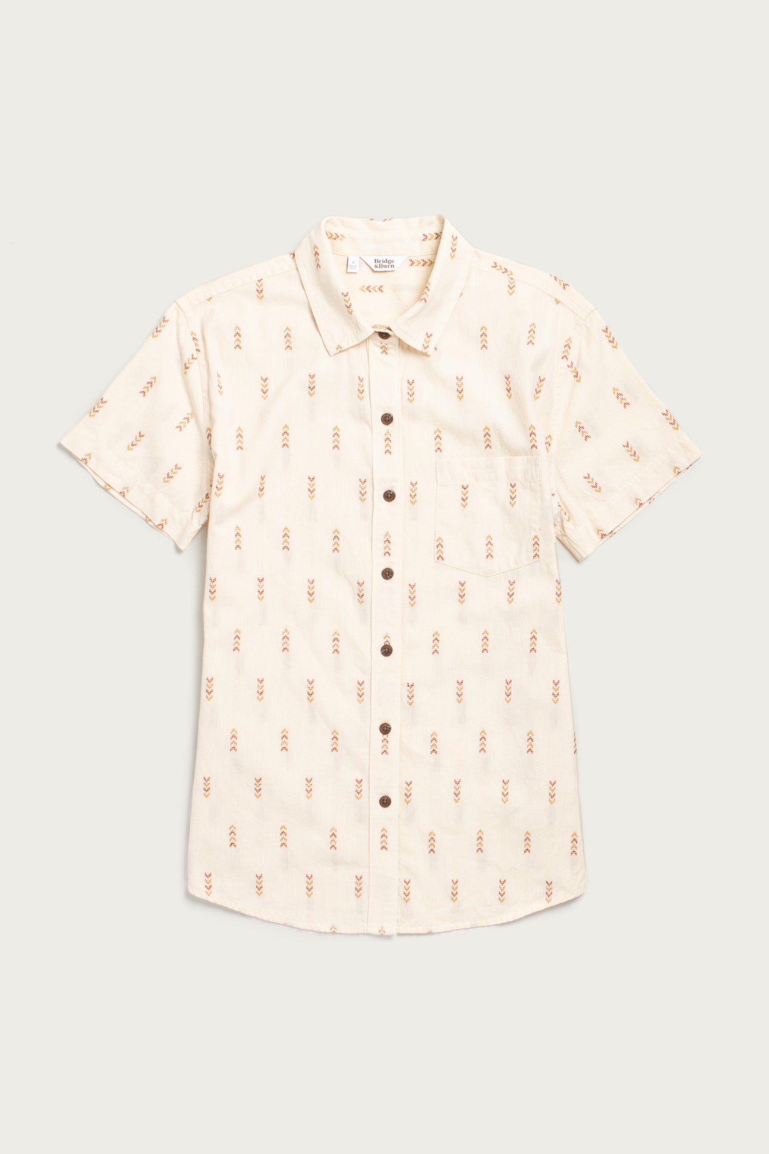 The Bridge & Burn Lana Shirt in Dune Chevron is a relaxed-fit, short sleeve button-up in light beige with a chest pocket and an all-over subtle red and brown chevron pattern.