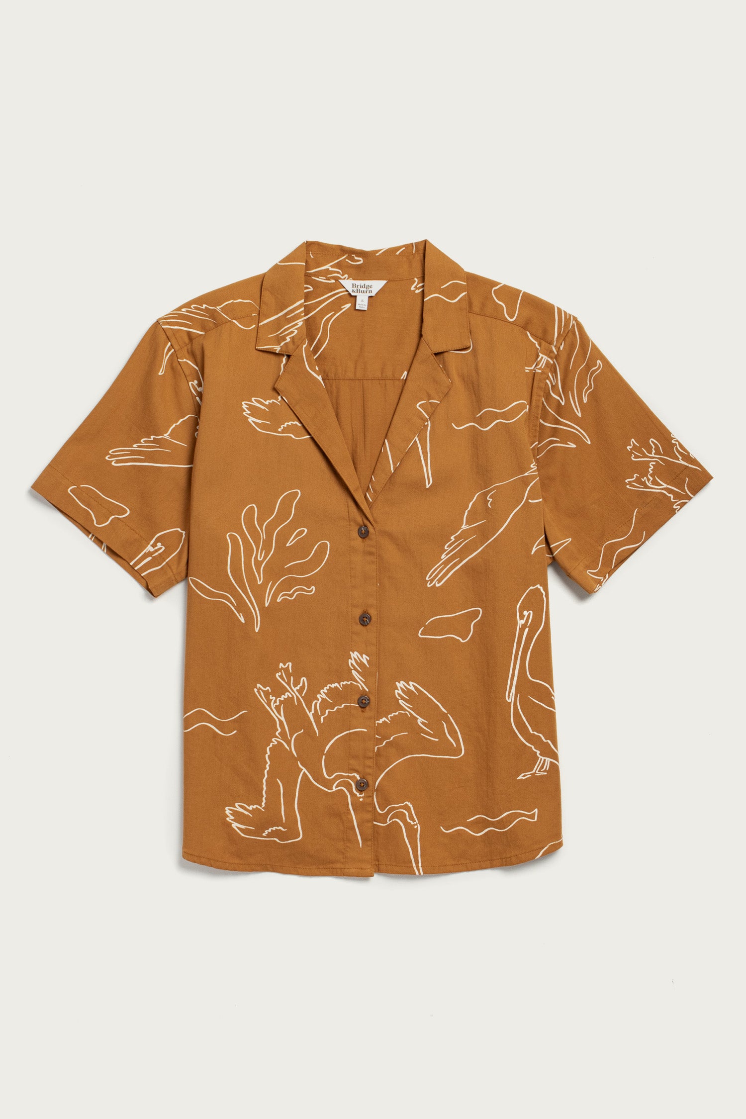 The Mila Cropped Shirt / Shoreflight by Bridge & Burn is a brown, short-sleeve, button-up with a notched collar and white abstract bird and plant print in organic cotton. The shirt is displayed flat against a light background.