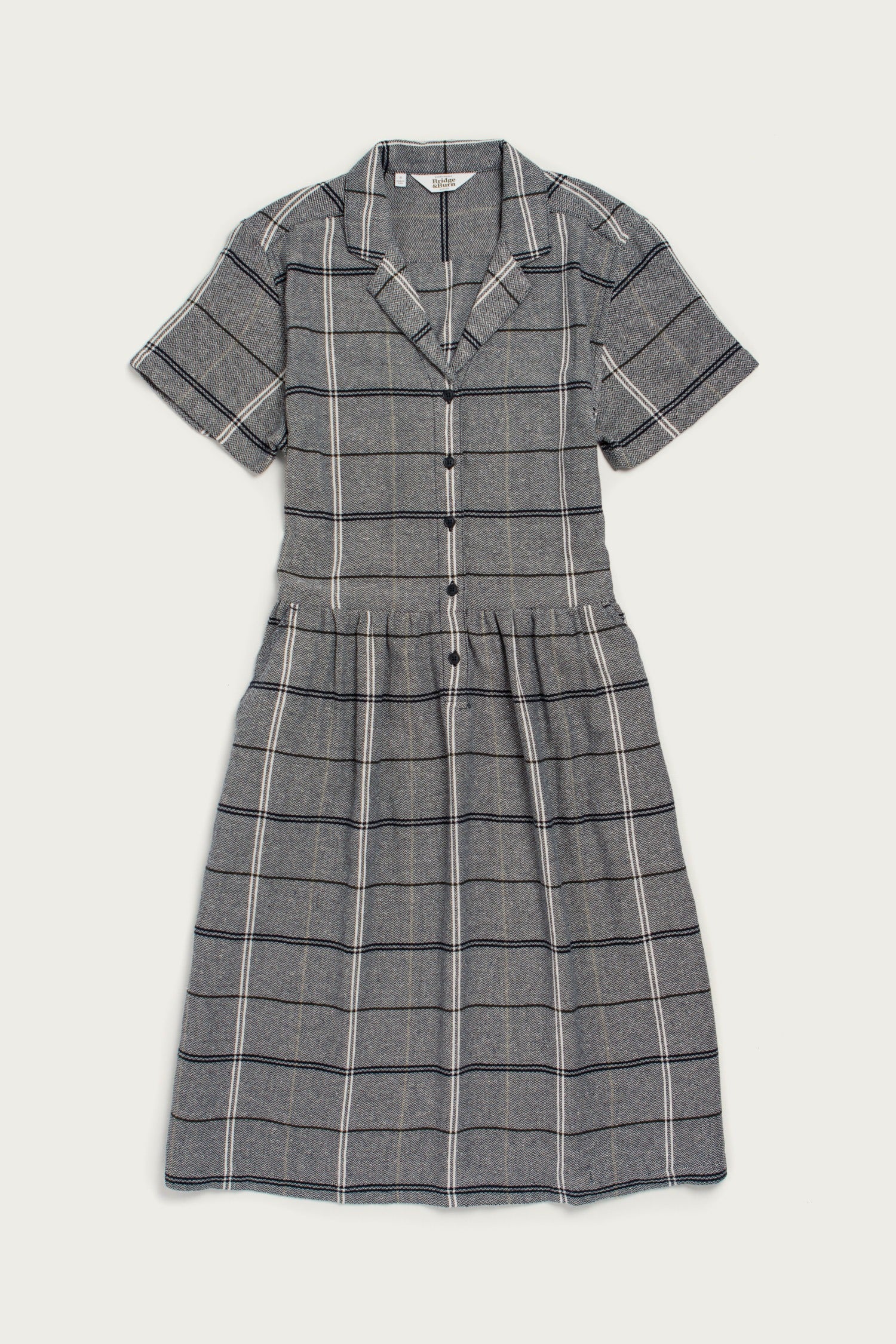 The Ruby Shirt Dress by Bridge & Burn features a collared neckline, front buttons, and fitted waist with a slightly flared knee-length skirt. Made from recycled cotton in Overcast Grid gray plaid, it’s perfect for effortless style.