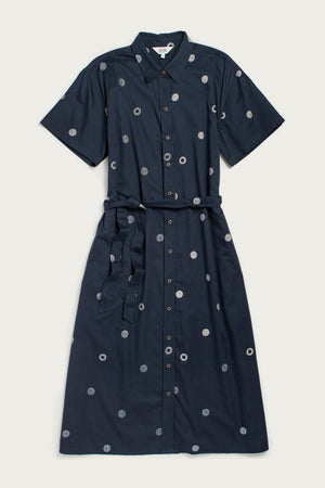 The Sylvie Dress / Lunar Polka by Bridge & Burn is a navy blue, oversized organic cotton shirtdress with short sleeves, a collar, a matching waist tie, and white circular patterns. It is shown laid flat on a plain background.