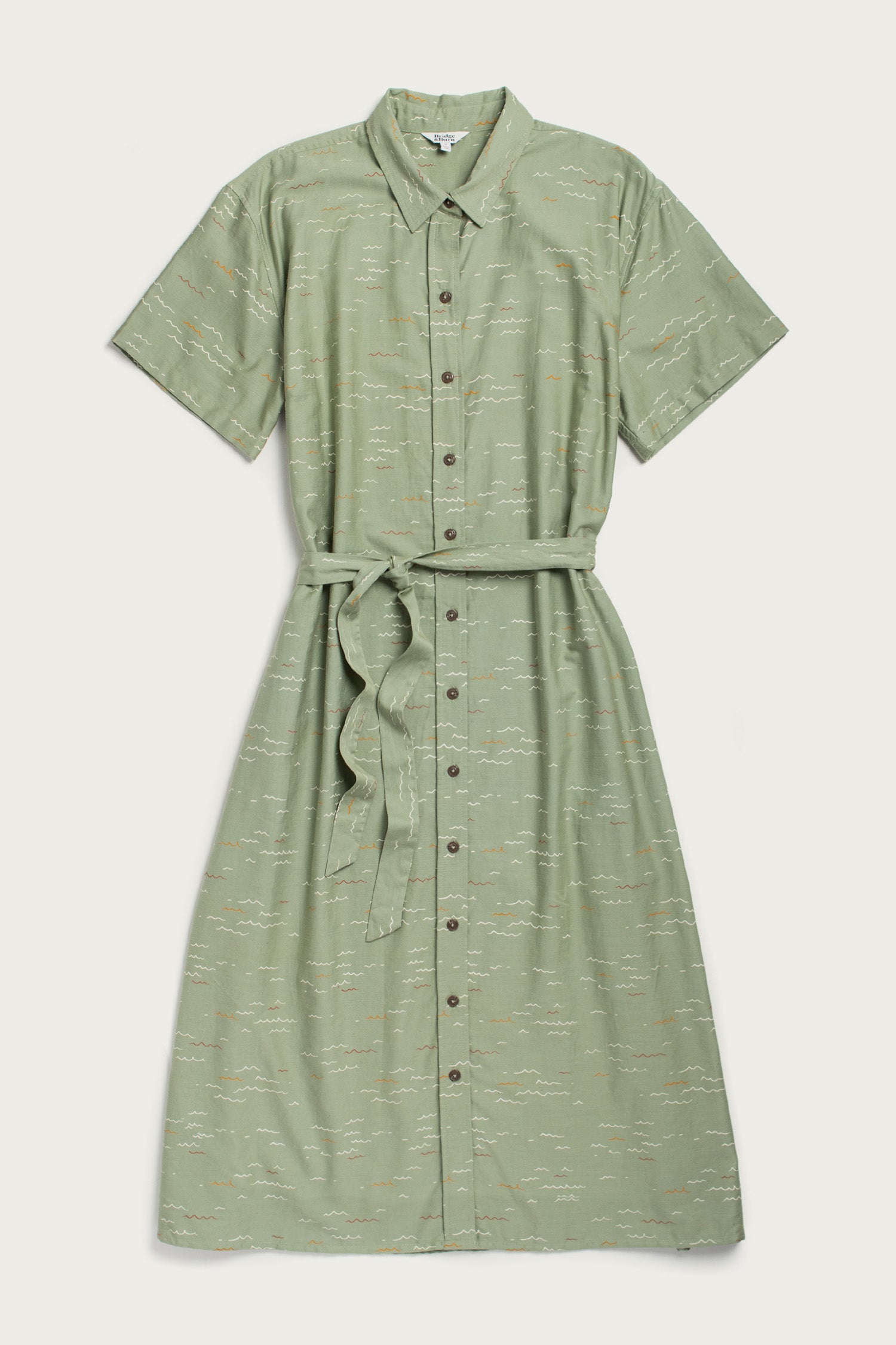 The Bridge & Burn Sylvie Dress in Teal Tide Waves is a light green, short-sleeve, collared shirtdress with a matching belt and subtle abstract print. Made from organic cotton, this relaxed midi dress is shown flat on a light background.