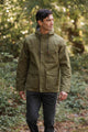 A man in the Bridge & Burn Marshall Jacket in olive waxed canvas stands outdoors among trees and greenery, wearing dark pants and looking to his left.