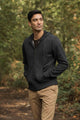 A man with short dark hair stands on a wooded path, wearing the Bridge & Burn Ridge Hoodie in Graphite—made from recycled fibers—and tan pants, hands in his pockets, surrounded by green trees and foliage.