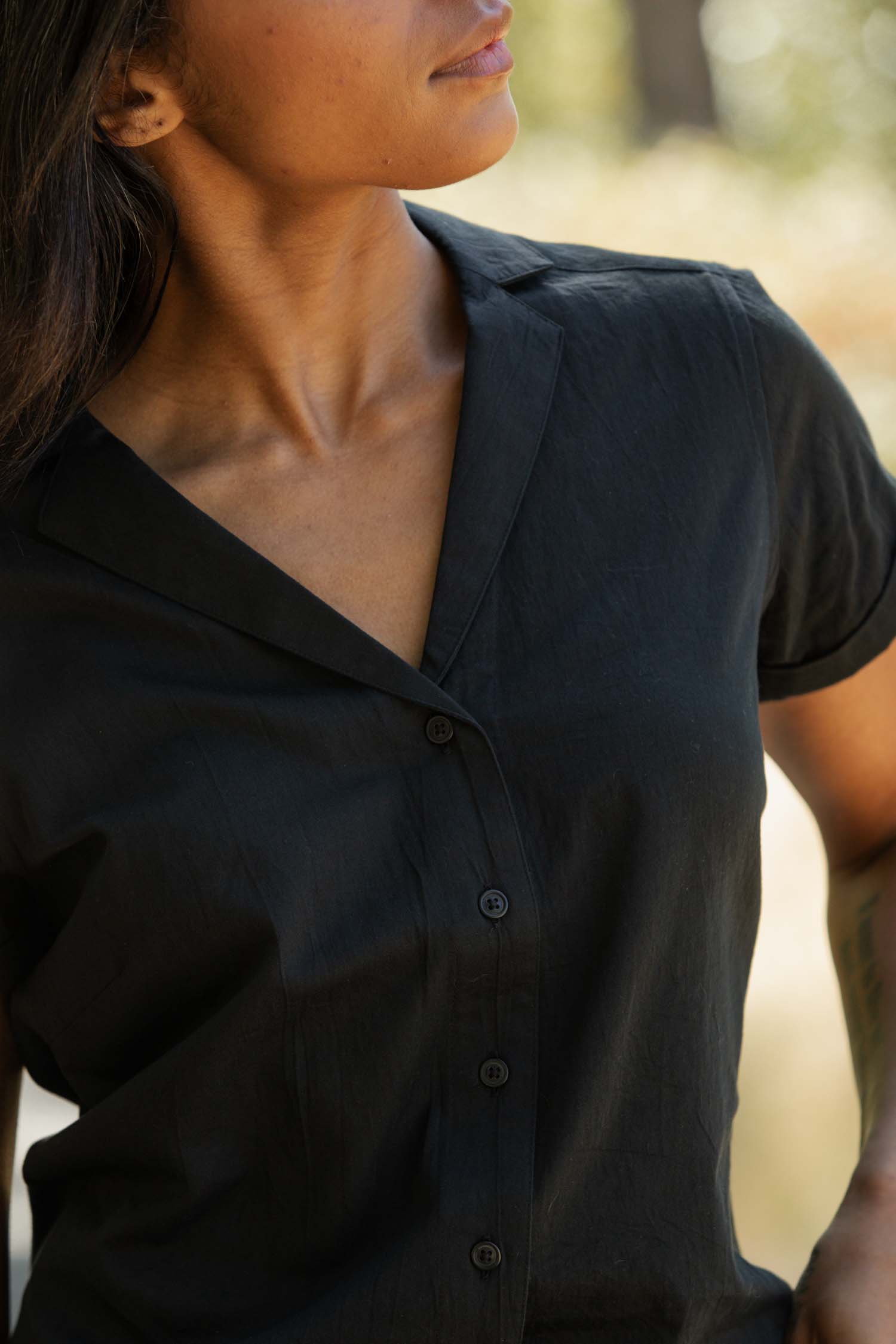 A person wearing the Bridge & Burn Innes Shirt in Black stands outdoors. The cropped image highlights their upper body and face, with sunlight illuminating their skin and the background.