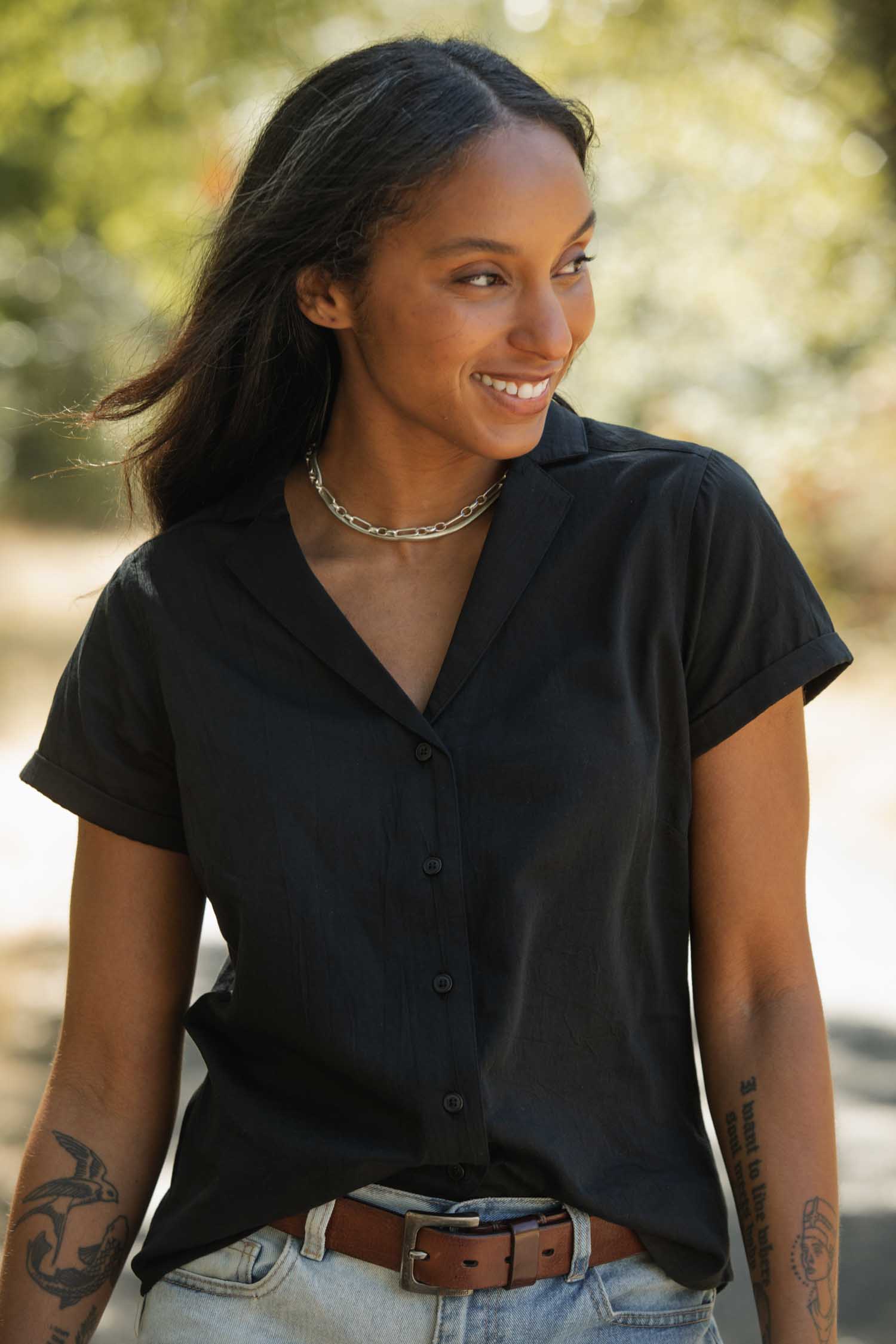 A woman with long dark hair smiles outdoors, wearing the Bridge & Burn Innes Shirt in black, light blue jeans, and a brown belt. She has forearm tattoos and a silver chain necklace.