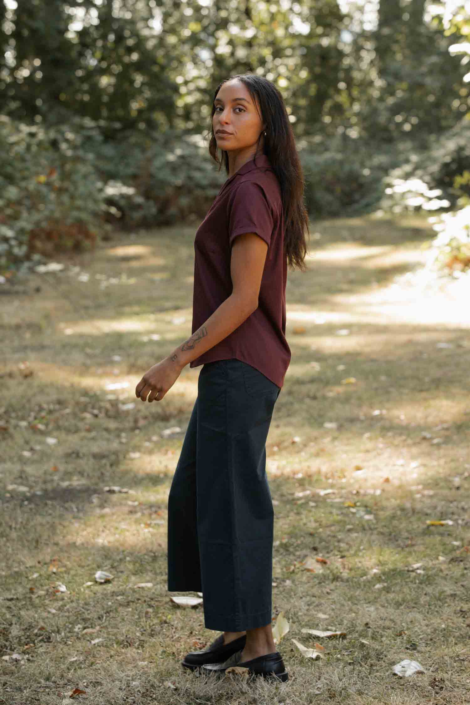 A woman with long dark hair stands on grass, wearing the Bridge & Burn Innes Shirt in Burgundy with wide-leg dark pants and black shoes. She looks to the side, surrounded by trees and sunlight.