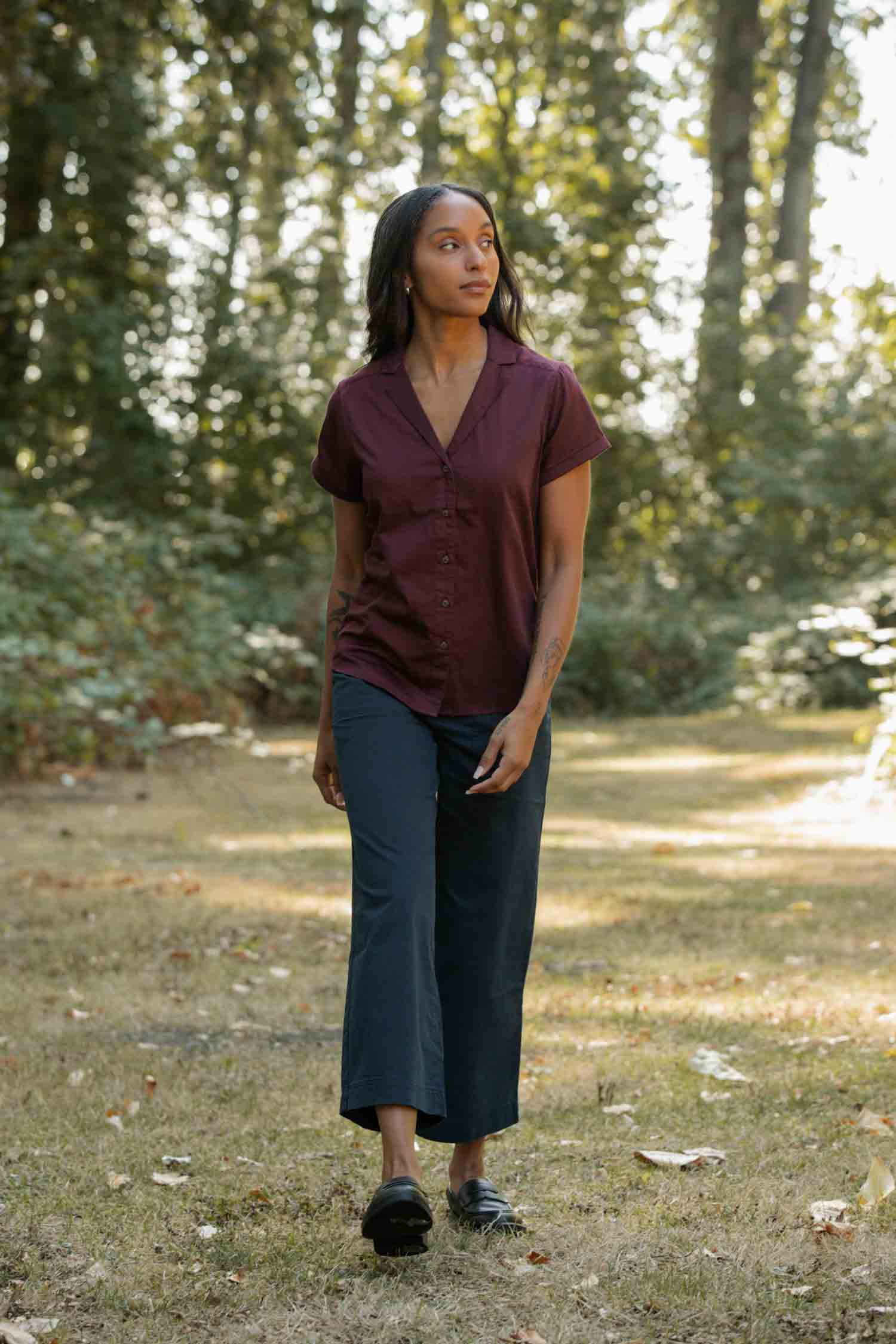 A woman with long hair stands on grass in sunlight, wearing the Bridge & Burn Innes Shirt in Burgundy, dark wide-leg pants, and black shoes. Trees and greenery appear in the background.