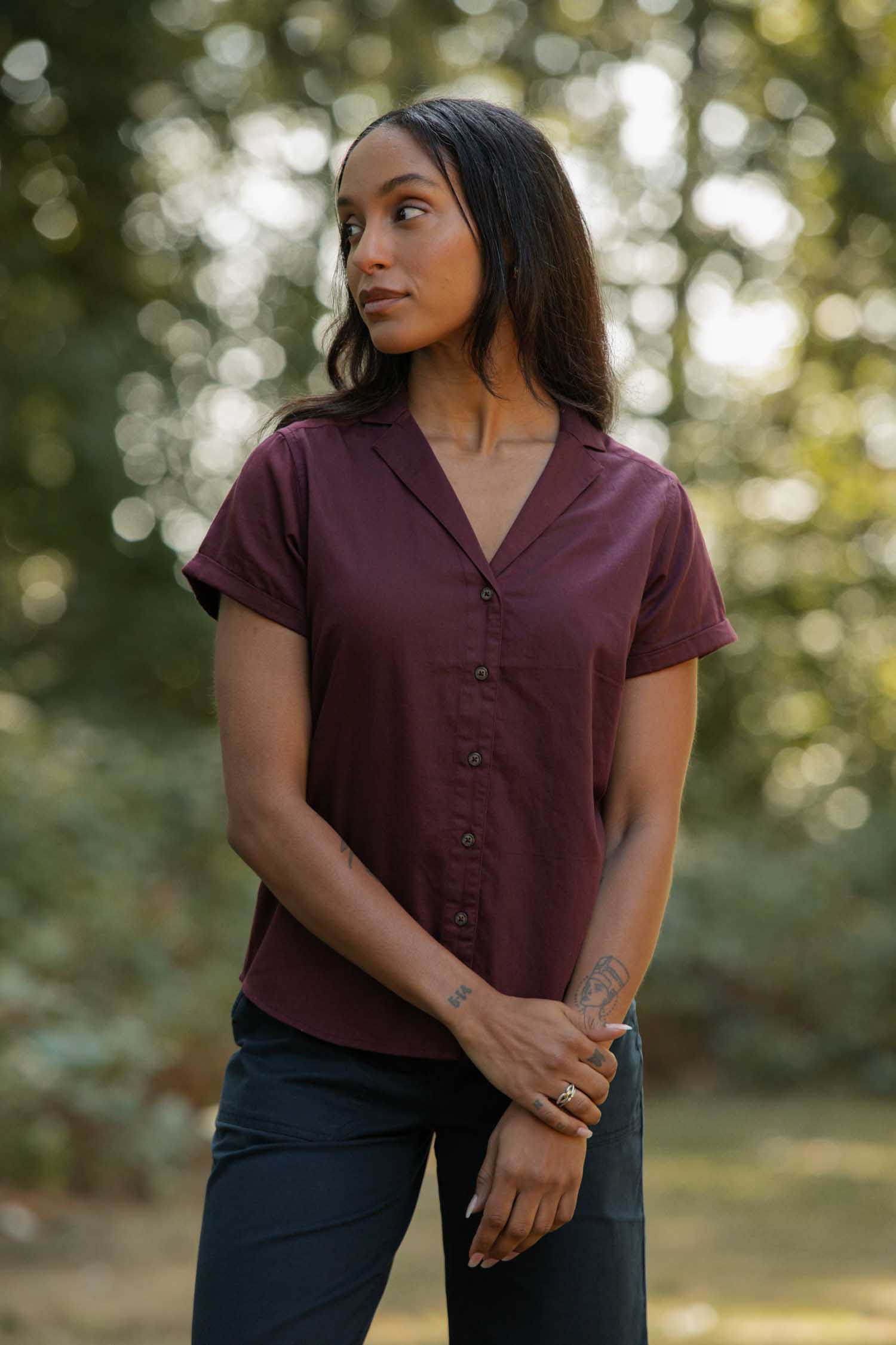 A woman stands outdoors before blurred trees, wearing the Bridge & Burn Innes Shirt in burgundy with dark pants. She looks to the side, arms loosely crossed in front of her.