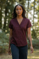 A woman with long dark hair and arm tattoos walks outdoors in the Innes Shirt in Burgundy by Bridge & Burn, paired with dark pants. Trees and greenery are blurred in the background.