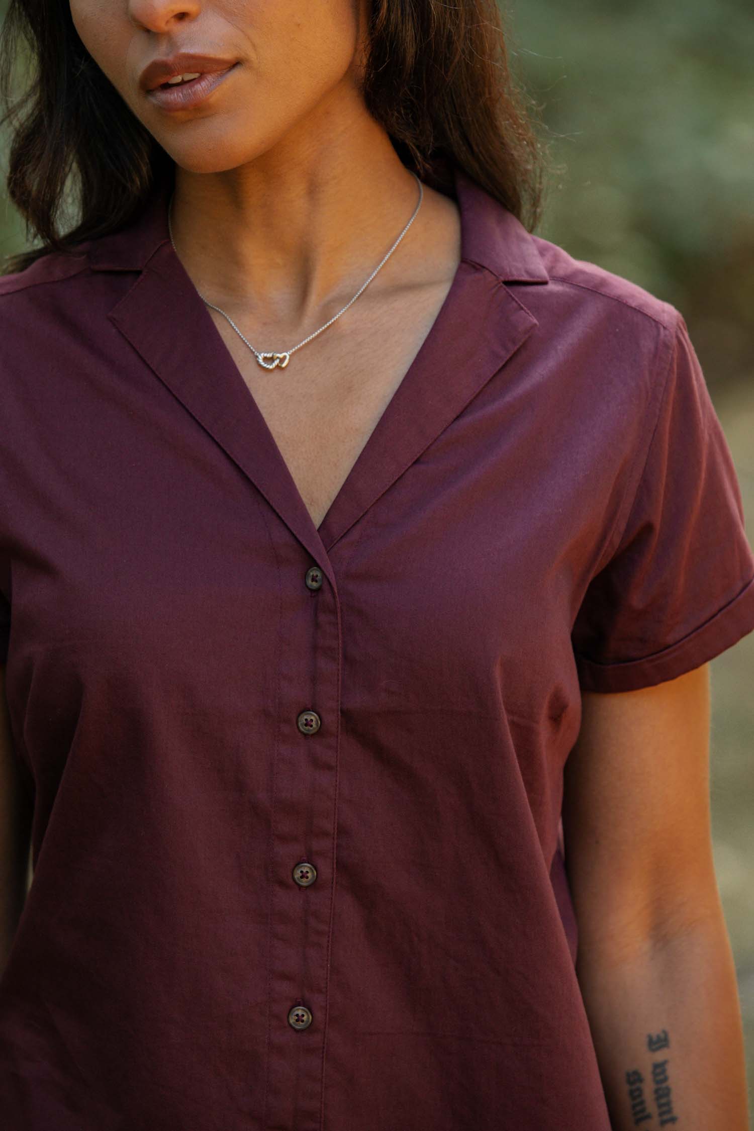 A woman stands outdoors wearing the Bridge & Burn Innes Shirt in burgundy, a relaxed fit button-up blouse. She pairs it with a silver infinity pendant necklace, with her tattooed forearm and part of her face visible.
