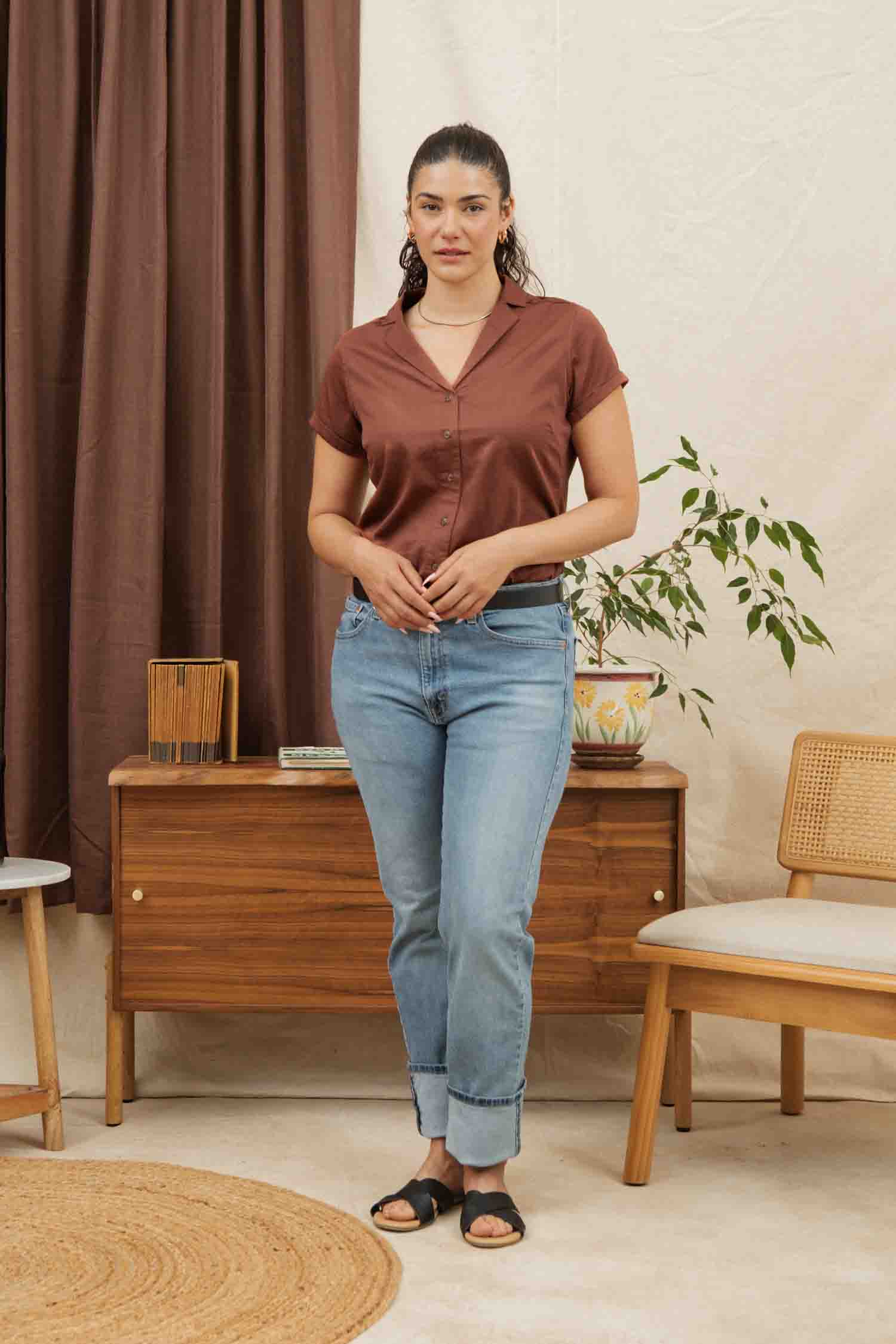 A woman with dark hair tied back stands indoors wearing the Bridge & Burn Innes Shirt in Copper, paired with light blue jeans and black sandals. She poses by a wooden cabinet, near a plant, decor, chairs, and a brown curtain.