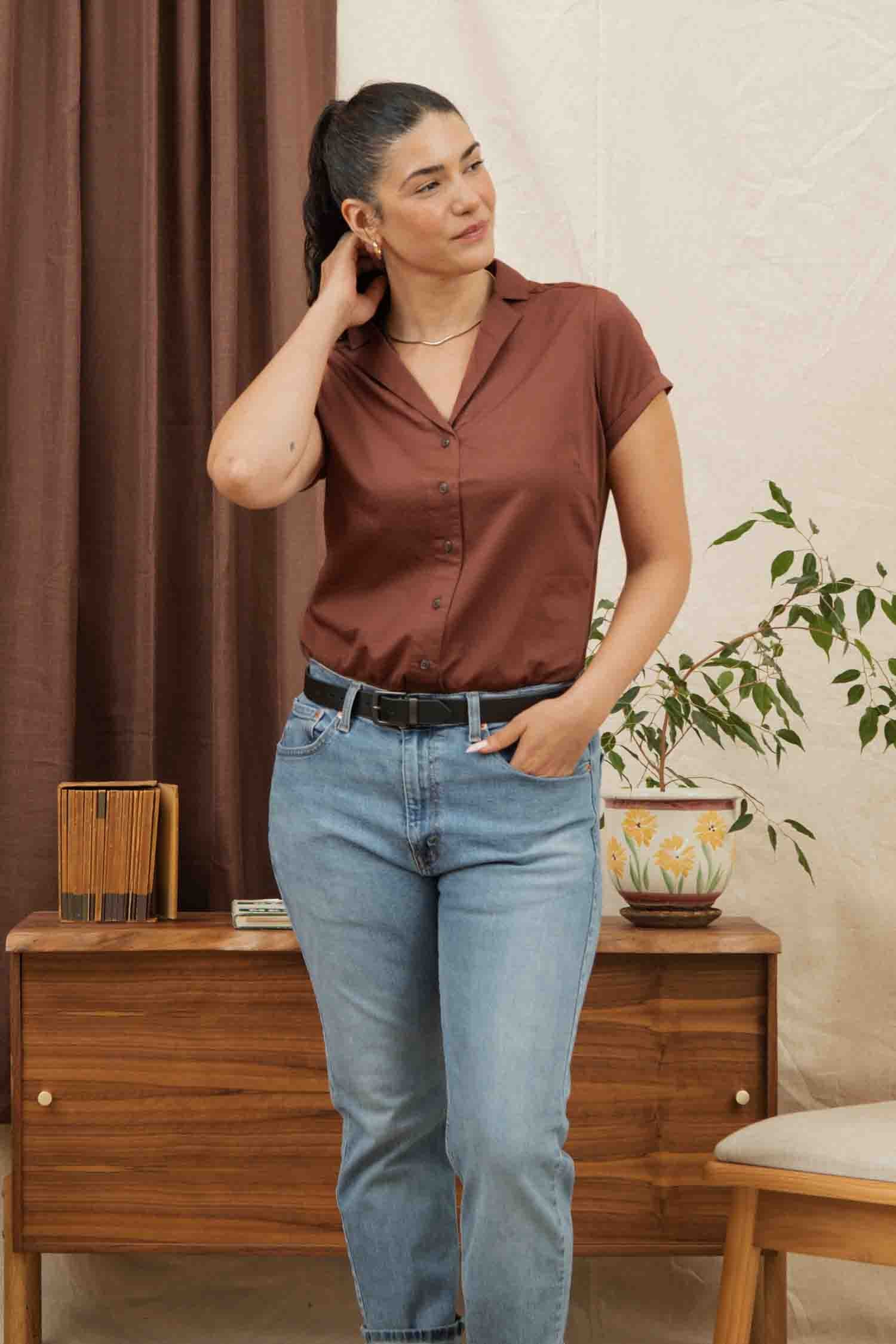 A woman with dark hair in a ponytail wears the Bridge & Burn Innes Shirt in Copper and light blue jeans, standing indoors by a wooden table with a potted plant, touching her neck and looking to the side.