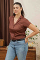 A woman with dark curly hair in a ponytail wears the Bridge & Burn Innes Shirt in Copper and blue jeans, standing indoors with one hand on her hip. A plant and vintage decor appear in the background.