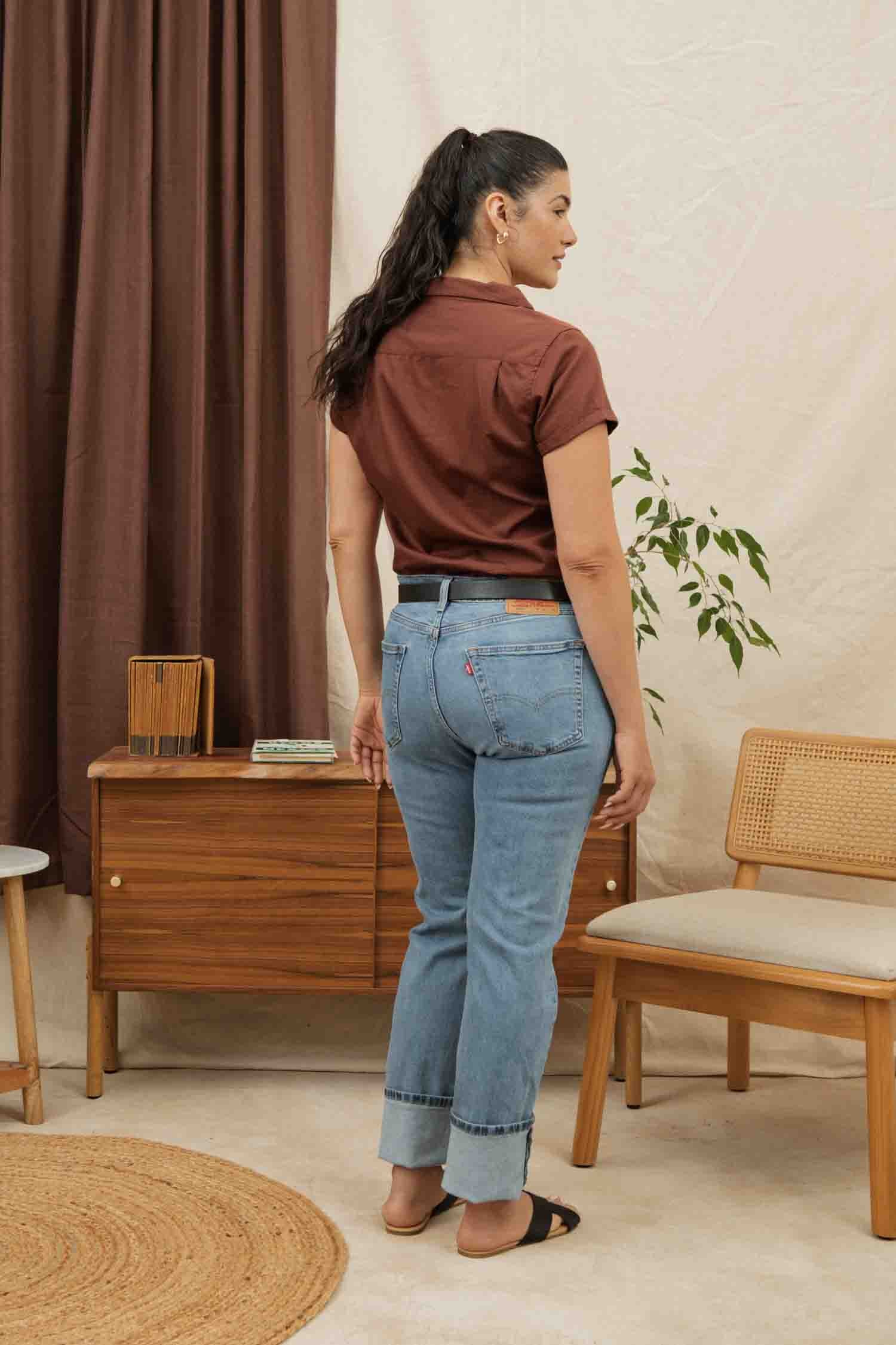A woman with long dark hair in a ponytail stands facing away, wearing the Bridge & Burn Innes Shirt in Copper, blue jeans, and black sandals. She is in a cozy room with wooden furniture, a plant, and neutral decor.