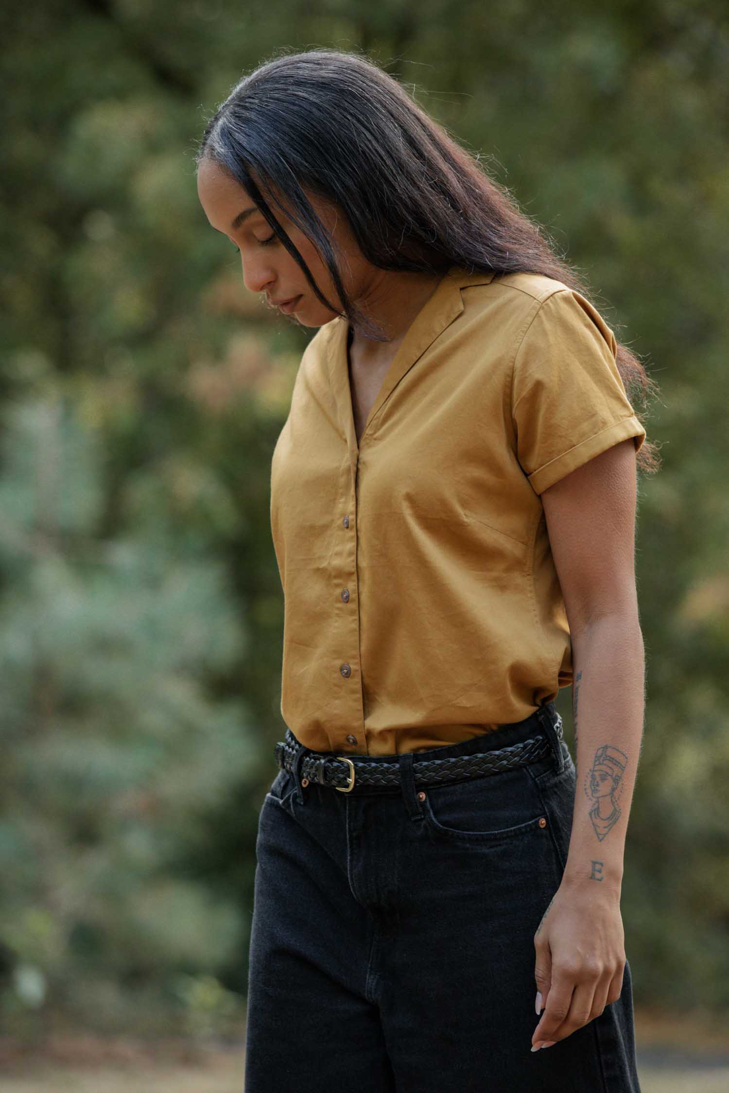 A woman with long dark hair wears the Bridge & Burn Innes Shirt in Gold and black pants outdoors, looking down. She has a tattoo on her left forearm, with blurred greenery in the background.