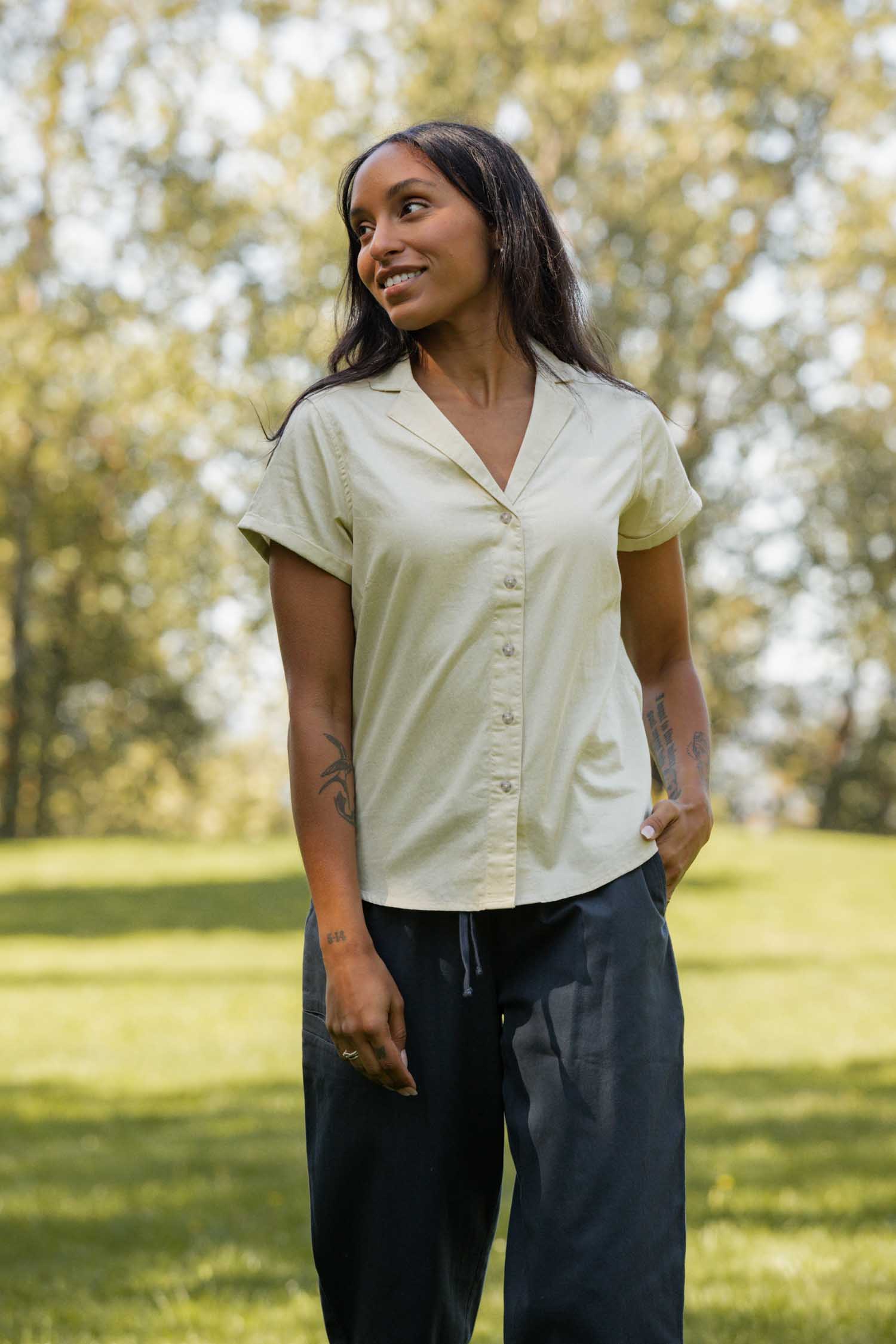 A woman with long dark hair stands on grass outdoors, smiling and looking to the side. She wears the Bridge & Burn Innes Shirt in Ivory, paired with dark pants. Trees and sunlight can be seen in the background.