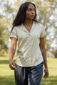 A woman with long dark hair and visible arm tattoos wears the Bridge & Burn Innes Shirt in Ivory, a relaxed fit short sleeve button-up, paired with dark pants as she walks outdoors on a sunny day with green grass and trees in the background.