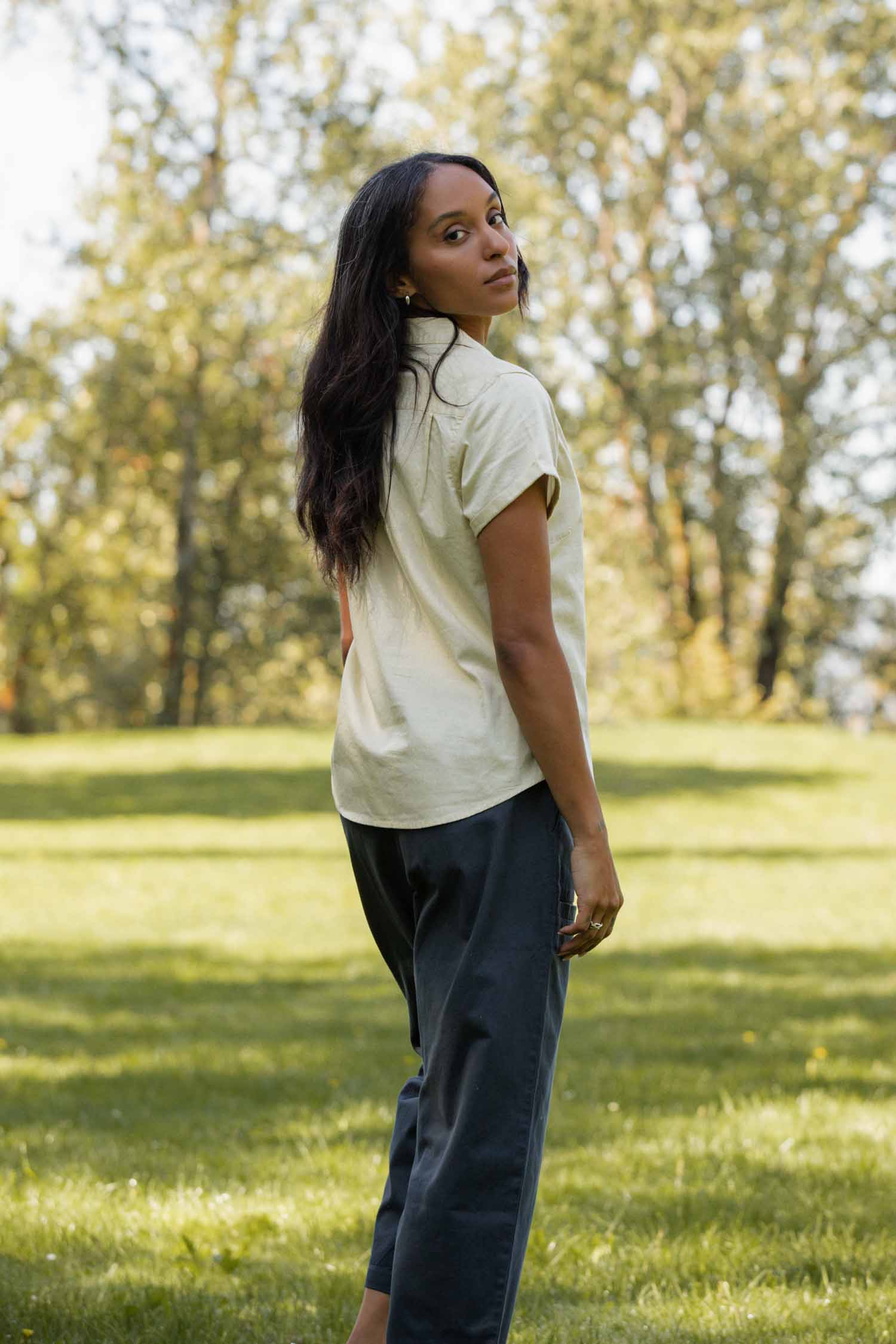 A woman with long dark hair stands on grass in a sunlit park, looking over her shoulder at the camera. She wears the Bridge & Burn Innes Shirt in Ivory and dark pants, with trees in the background.