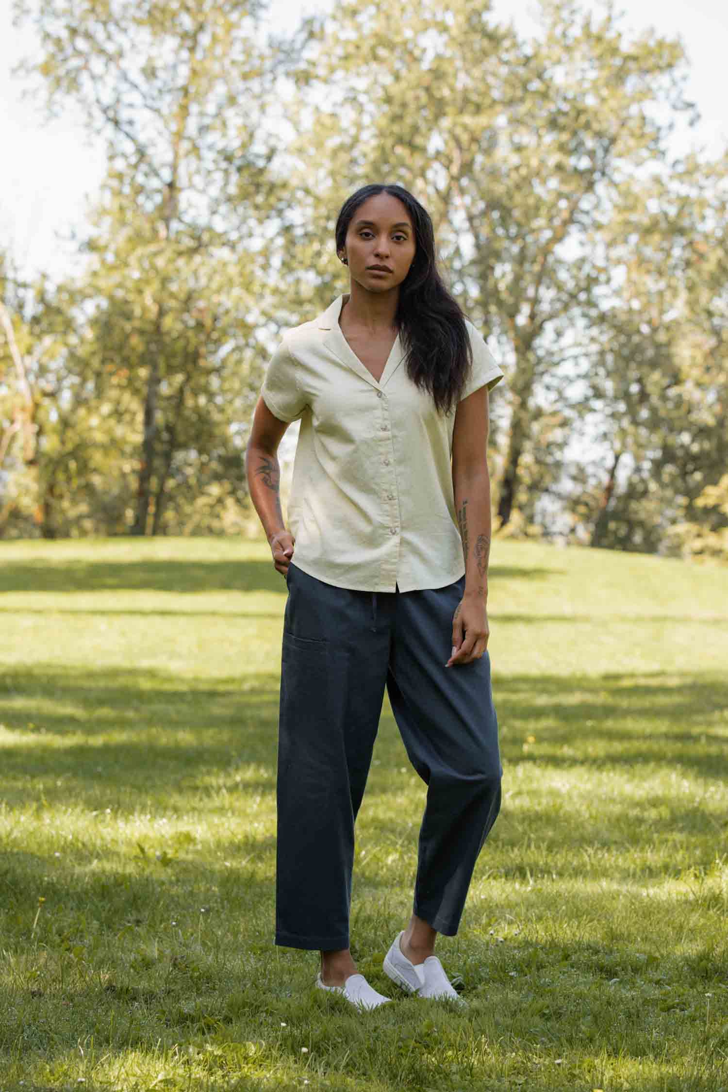 A woman stands on grass in a park, wearing the Bridge & Burn Innes Shirt in Ivory—a relaxed fit, short sleeve button-up blouse—paired with dark loose-fitting pants and white shoes. Trees and sunlight create a serene, natural backdrop.