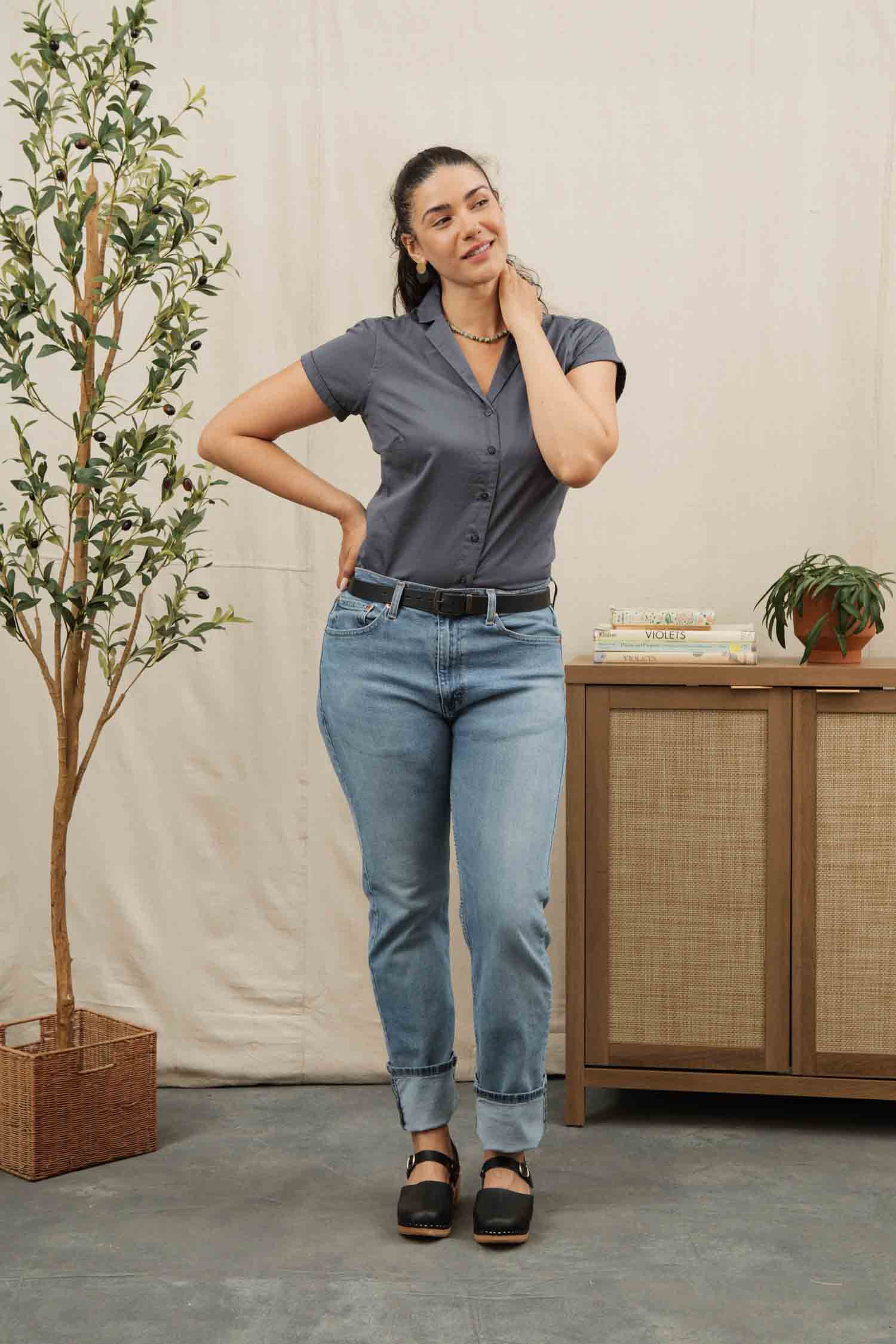 A woman stands indoors by a leafy plant and wooden sideboard, smiling in the Bridge & Burn Innes Shirt in Light Navy, styled with light blue jeans and black shoes, her hand resting on her neck.