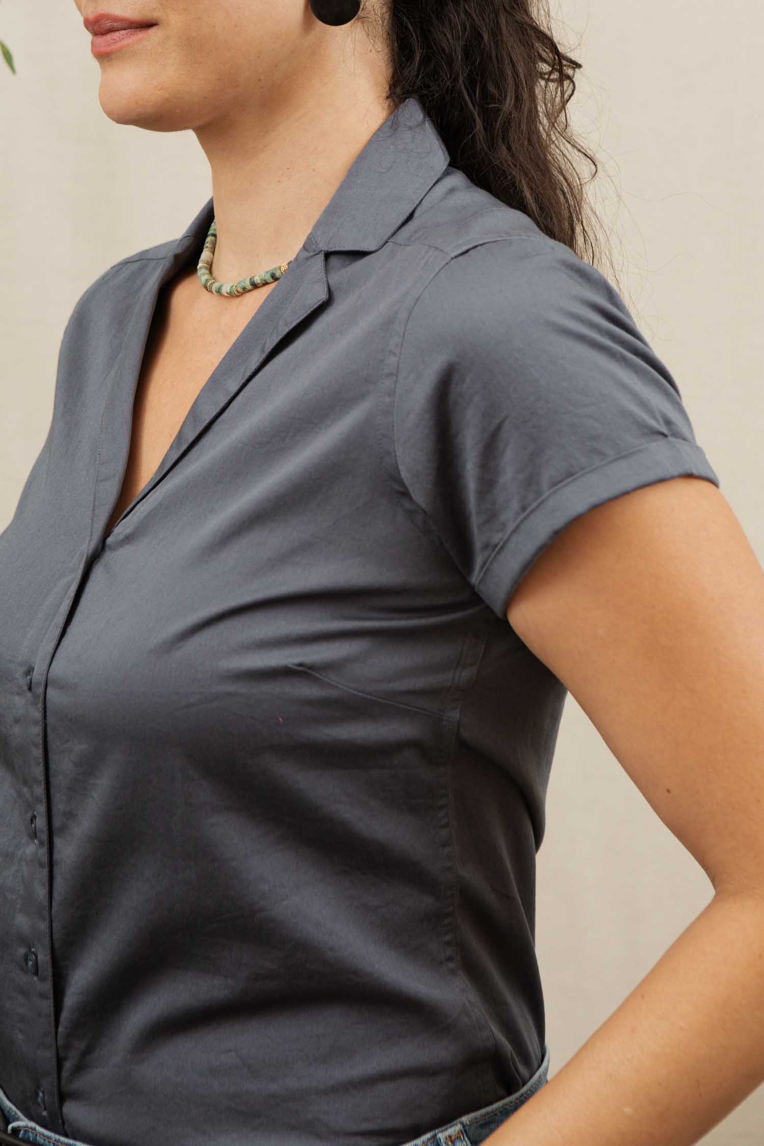 A woman wears the Bridge & Burn Innes Shirt in light navy, a relaxed fit button-up short sleeve blouse, paired with a beaded necklace. She stands with her hand on her hip, upper body visible, facing left.