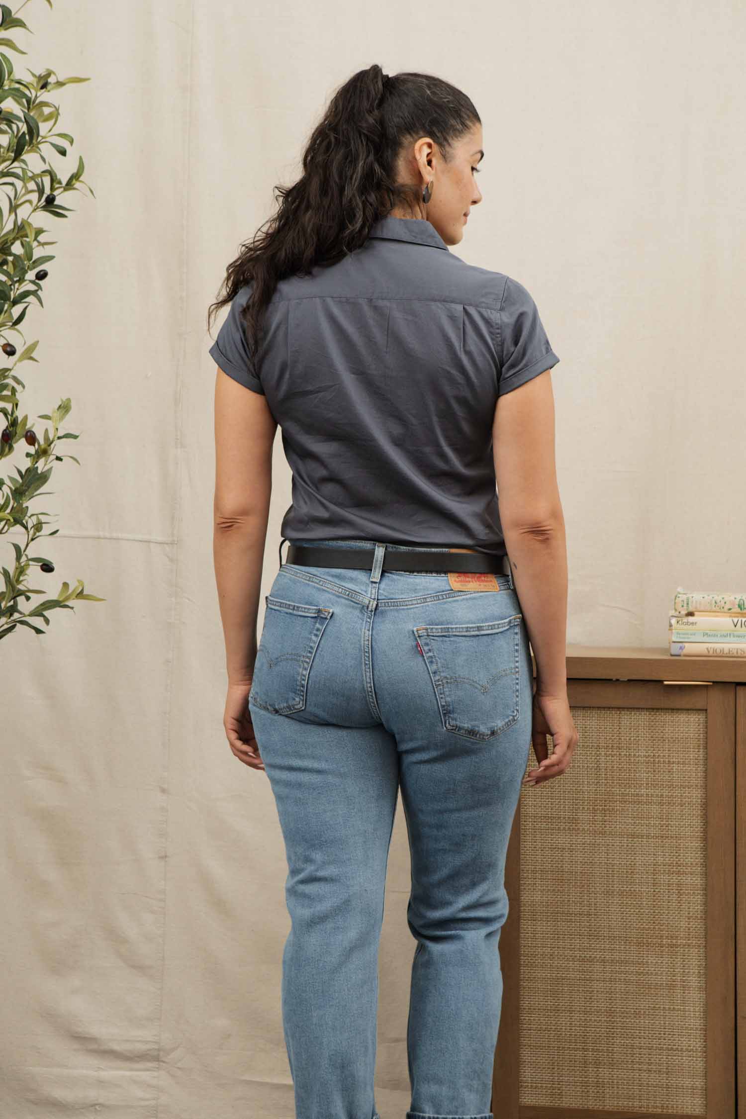 A woman with long dark hair in a ponytail stands indoors by a wooden table with books and a plant, wearing the Bridge & Burn Innes Shirt in Light Navy and light blue jeans.