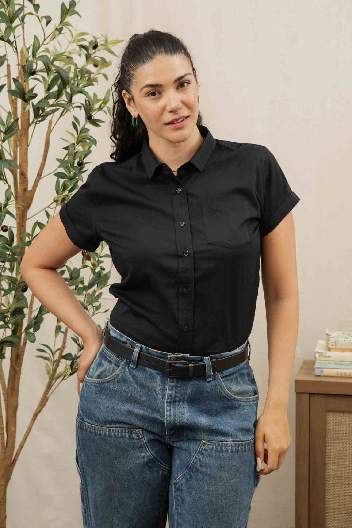 A woman with dark hair in a ponytail wears the Bridge & Burn Bea Shirt in Black with blue jeans. She stands indoors, one hand on her hip, next to a plant and a book-stacked table.
