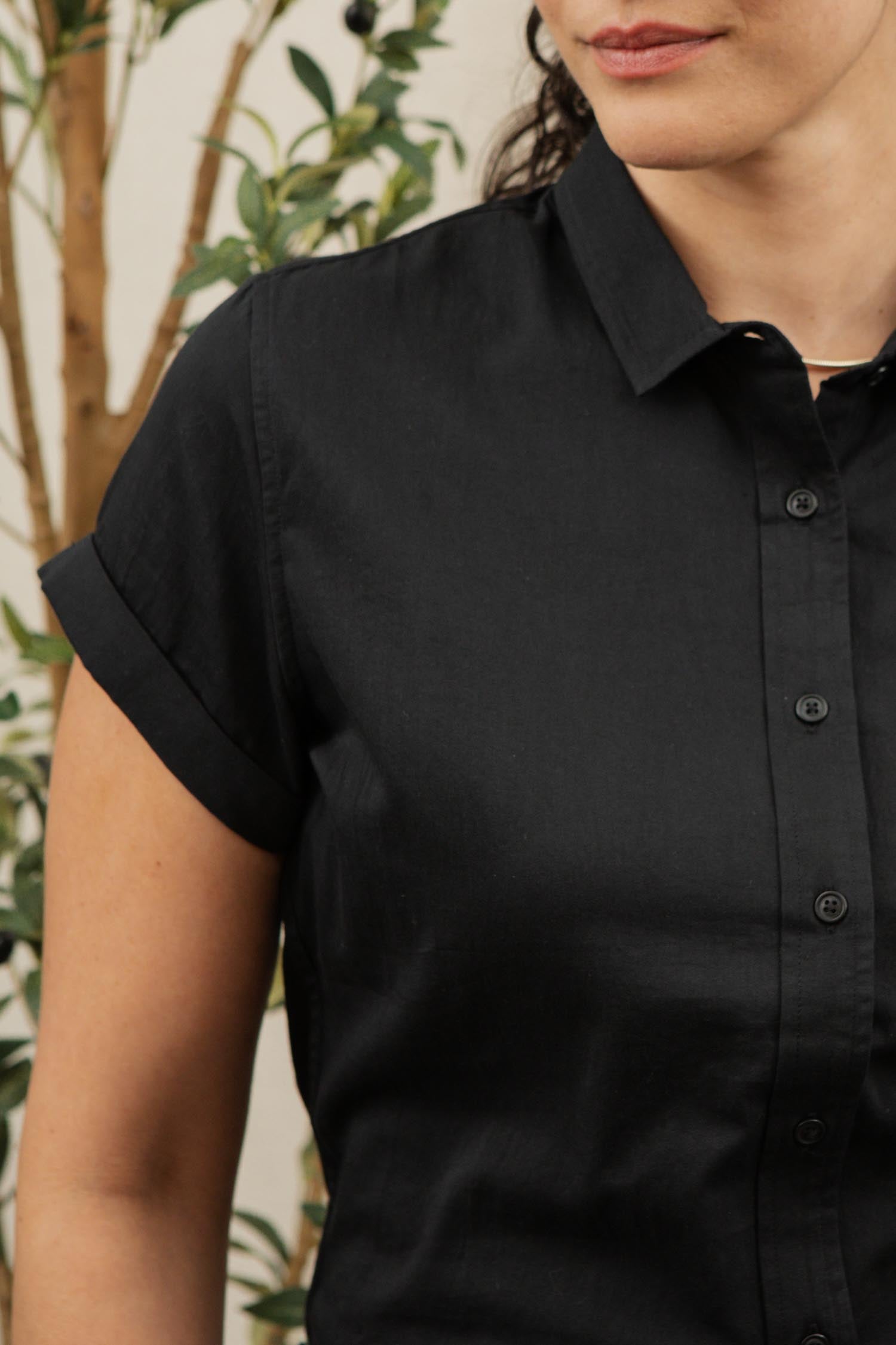 A woman models the Bridge & Burn Bea Shirt in black—a classic tailored, short-sleeve button-down—as she stands before a blurred plant background. Only her upper body and part of her face are visible.